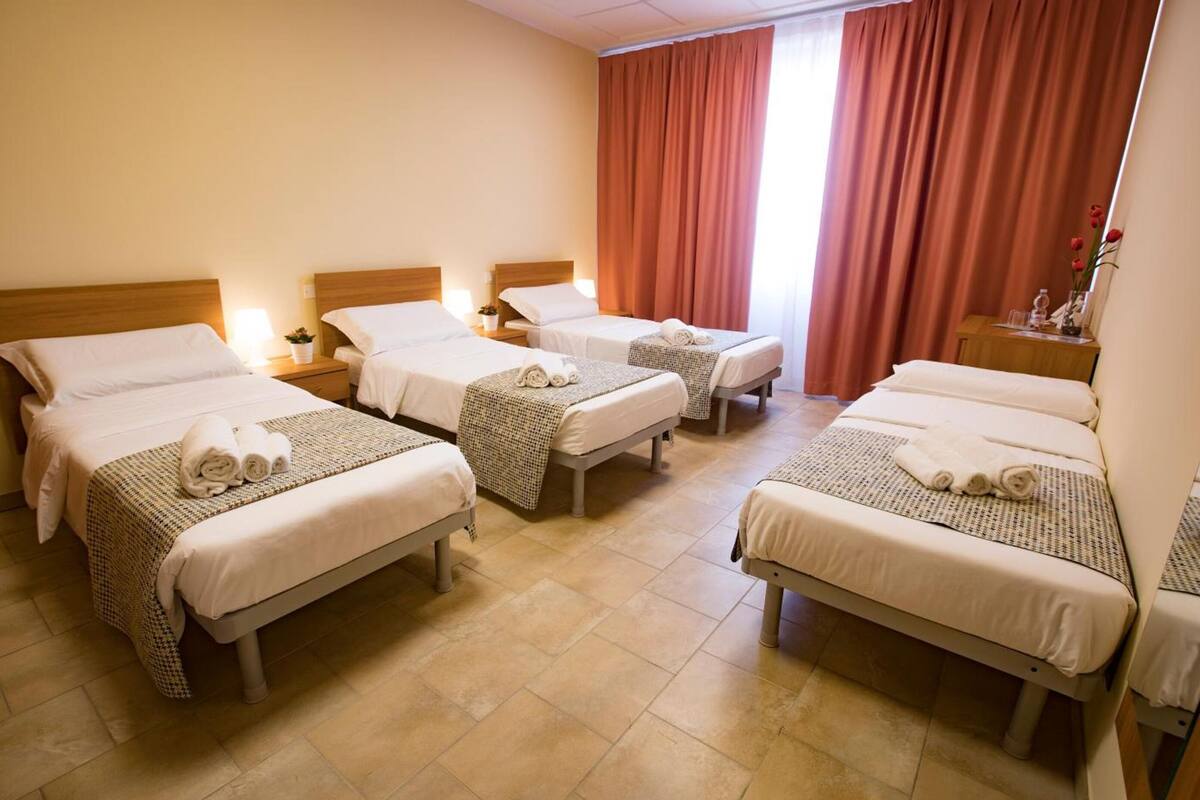 Une chambre d’hôtel avec quatre lits simples, chacun agrémenté d’une serviette soigneusement pliée, d’une table de chevet avec une lampe et d’une petite plante.
