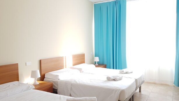 A hotel room with two beds, a nightstand with a lamp, and a large window with blue curtains. - Santa Teresa House (Verona)