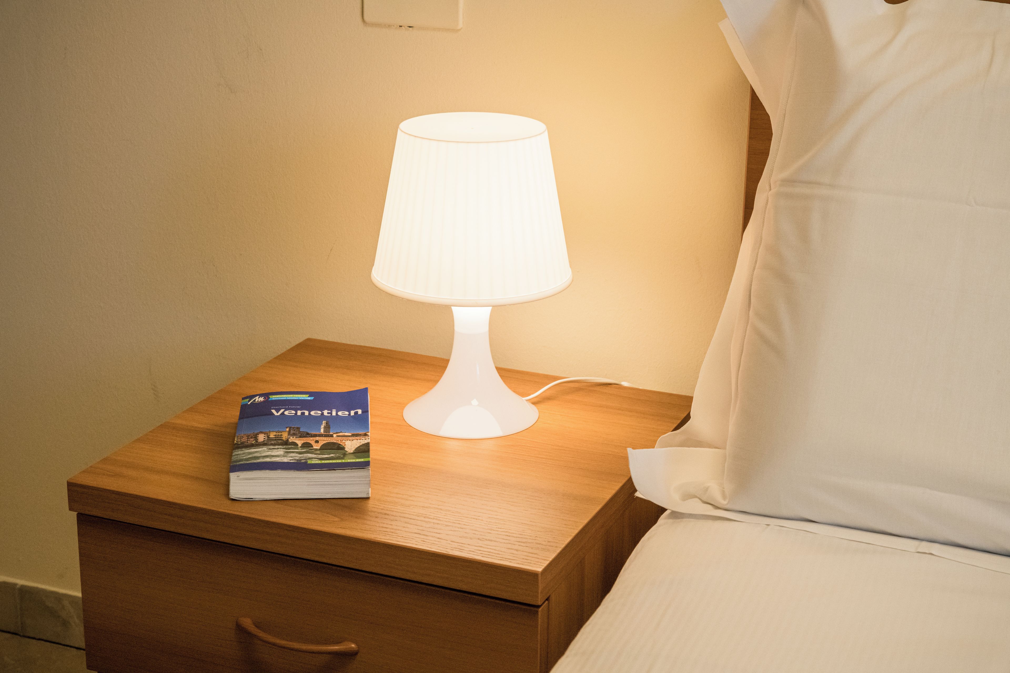 A bedside table with a lamp, a book titled 'Venetien', and a pillow.