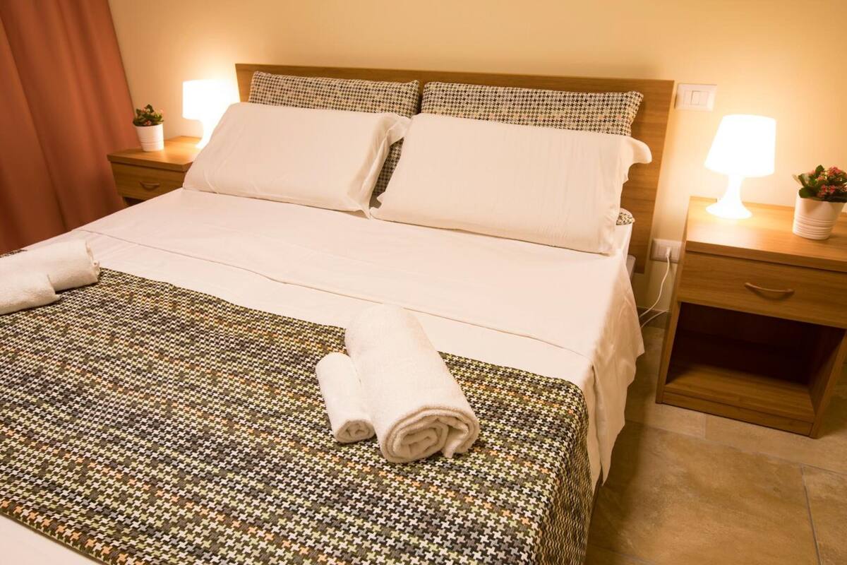 Un lit bien fait, avec du linge de lit blanc, une couvre-lit à motifs et deux serviettes enroulées posées sur le lit. Il y a deux tables de chevet, chacune avec une lampe, et une plante en pot sur l’une d’elles.