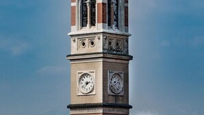 Un campanar amb un pinacle apuntat, dues cares de rellotge visibles i la inscripció "PUIUT HORO" a la torre.