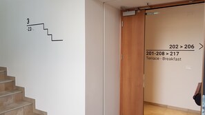 A hotel interior with a staircase marked with numbers, a sliding door with room numbers, and a sign indicating a terrace and breakfast area. - Santa Teresa House (Verona)