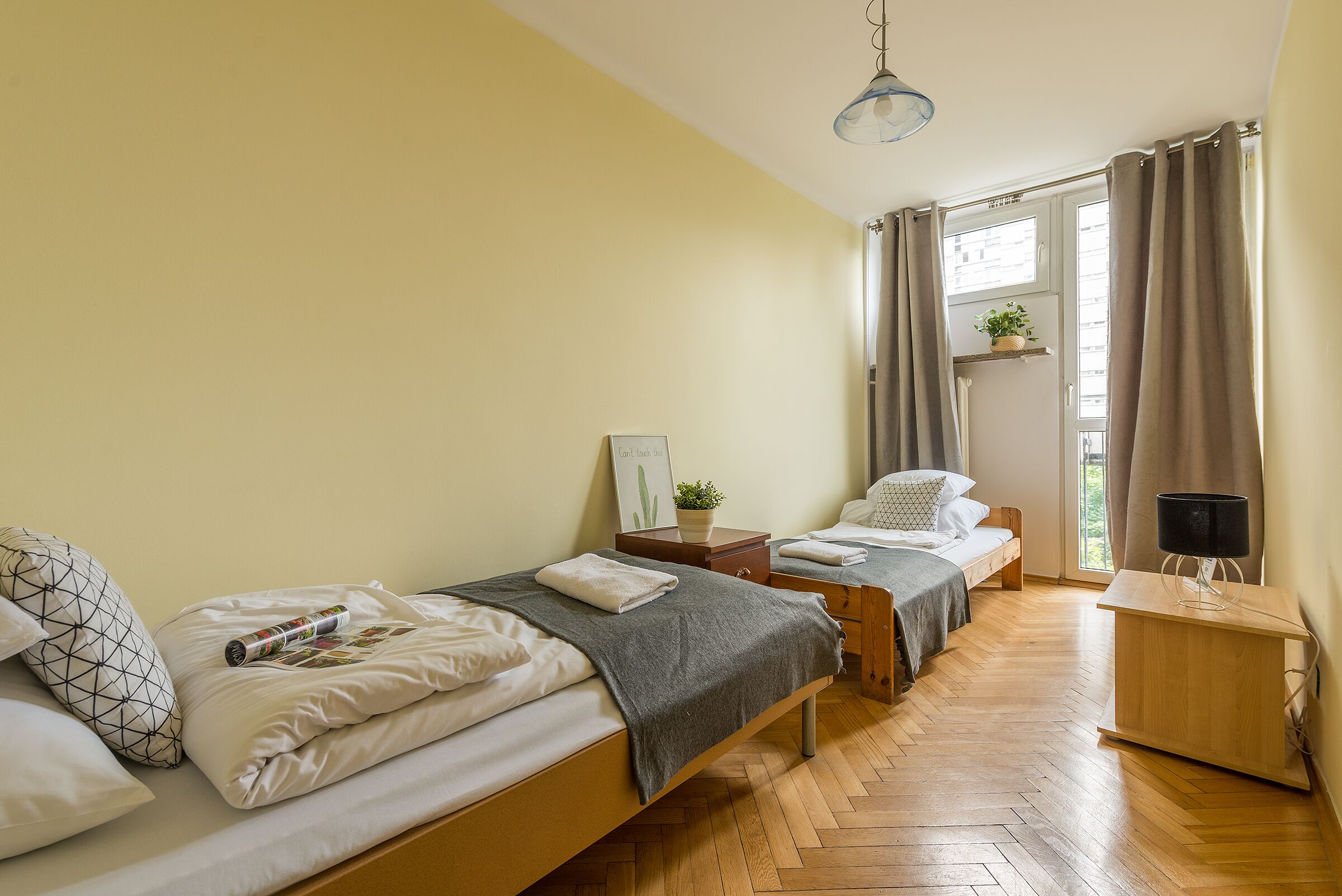 A bedroom with two beds, a wooden floor, a bedside table with a lamp, a window with curtains, and a small plant on the window sill.