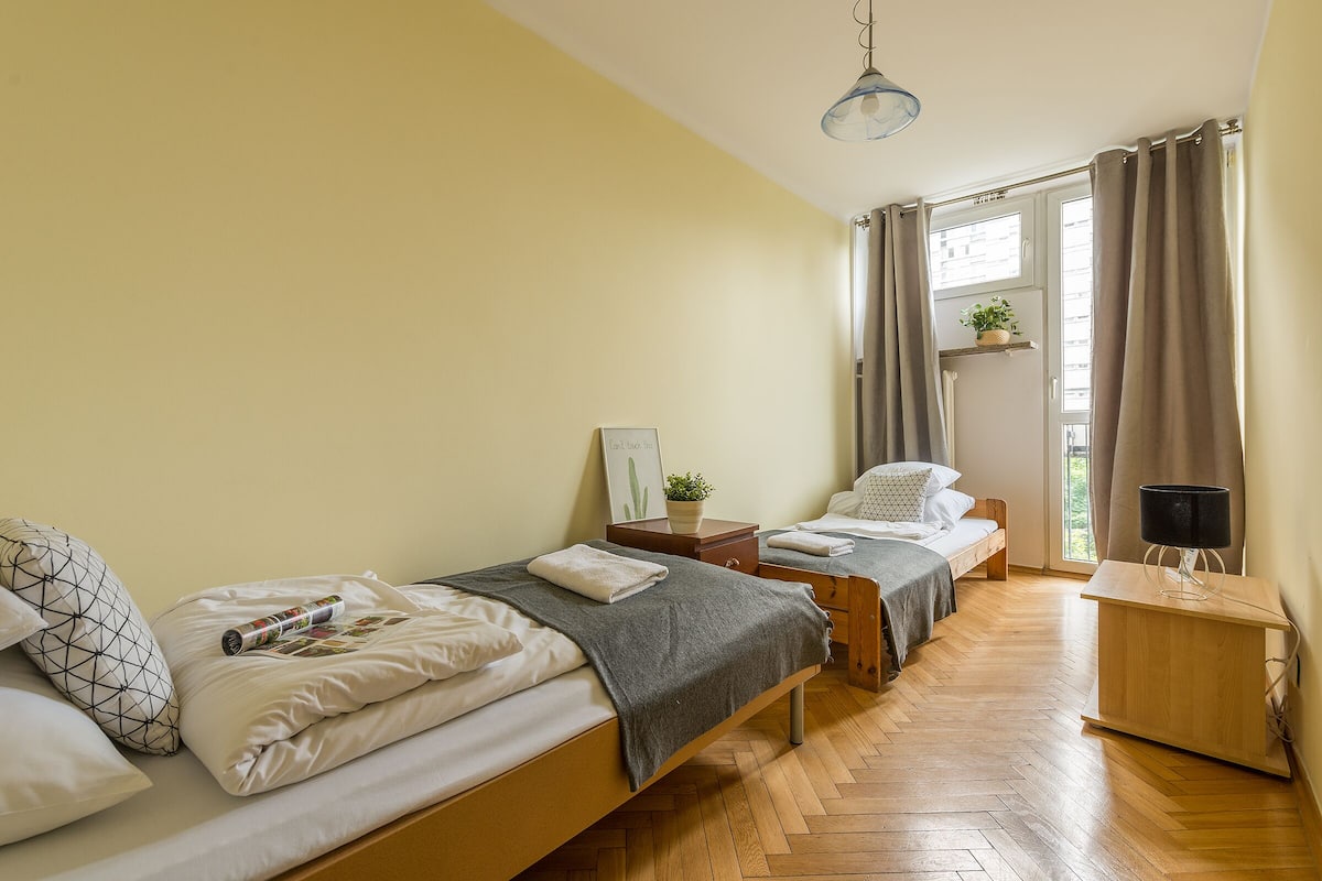 A bedroom with two beds, a wooden floor, a bedside table with a lamp, a window with curtains, and a small plant on the window sill.