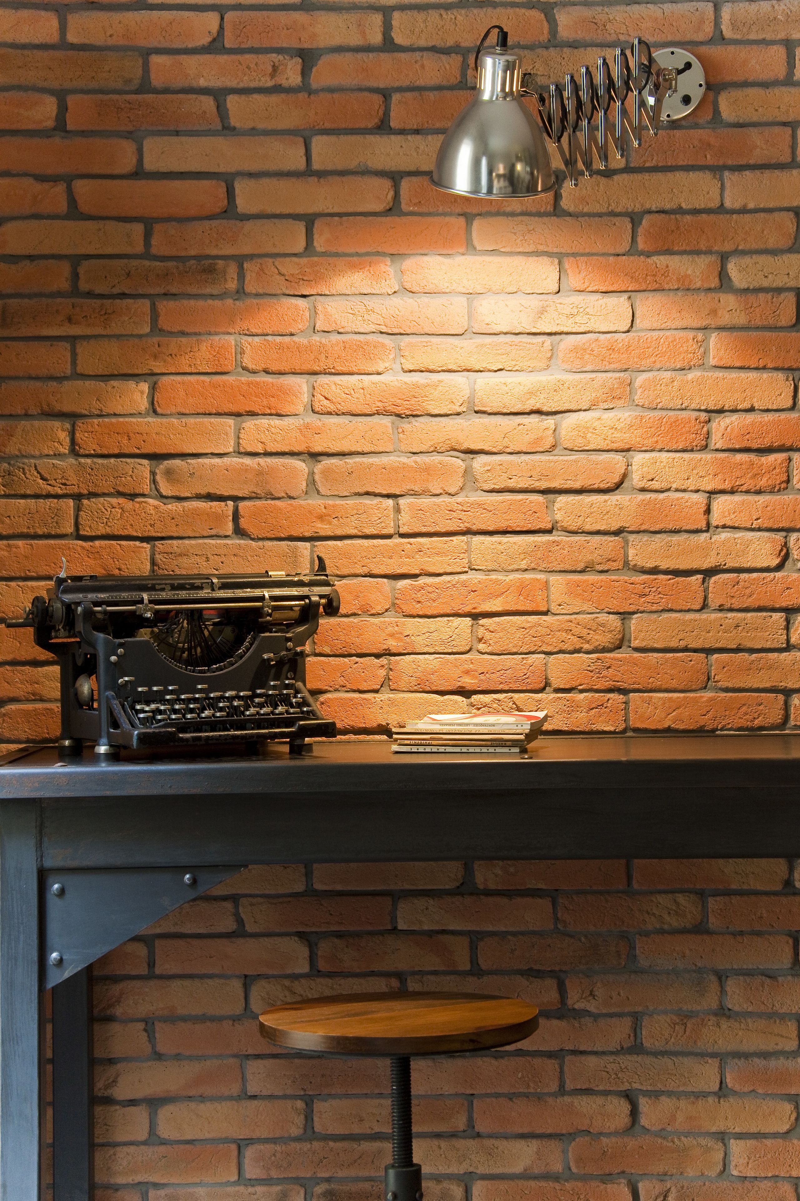 Une machine à écrire ancienne posée sur un bureau, avec une lampe et des livres, le tout adossé à un mur de briques.
