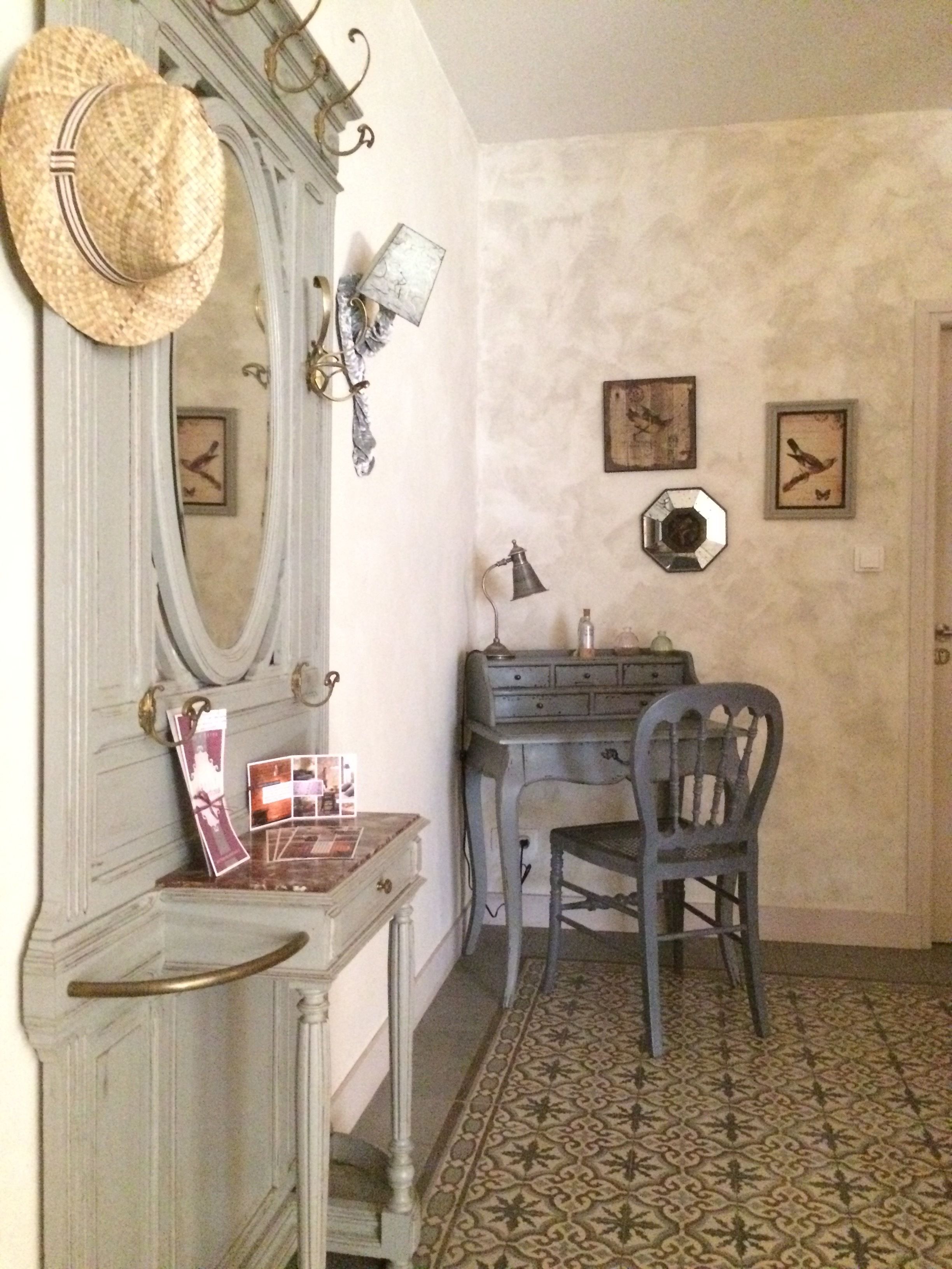 A room with a vintage-style dresser, a mirror, a hat on the wall, a desk with a lamp, and a chair.