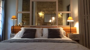 Un lit double avec une tête de lit, deux lampes de chevet, une table de chevet, un miroir et un mur en pierre derrière le lit.