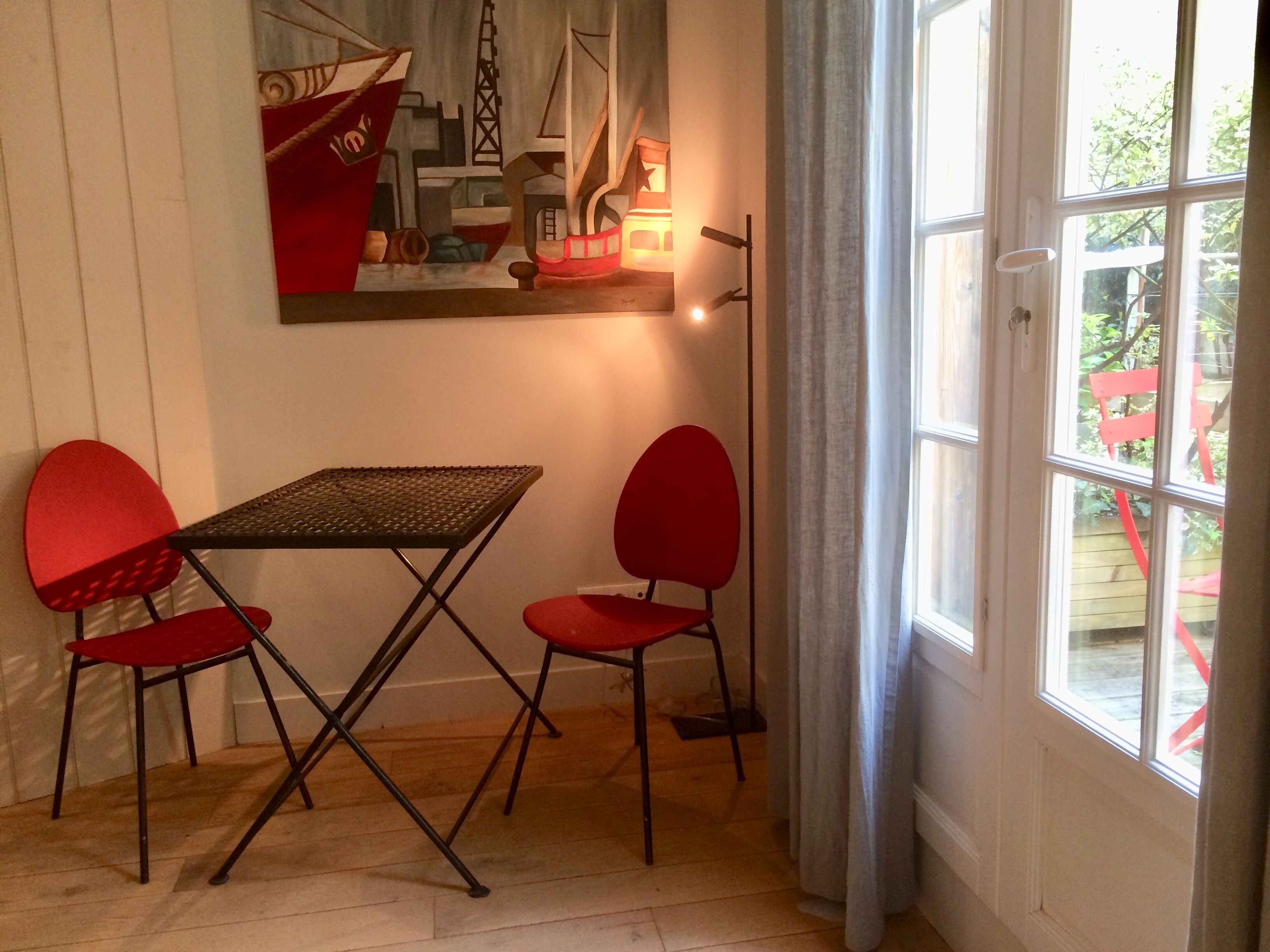 A room with a table and two red chairs, a painting on the wall, and a window with curtains.