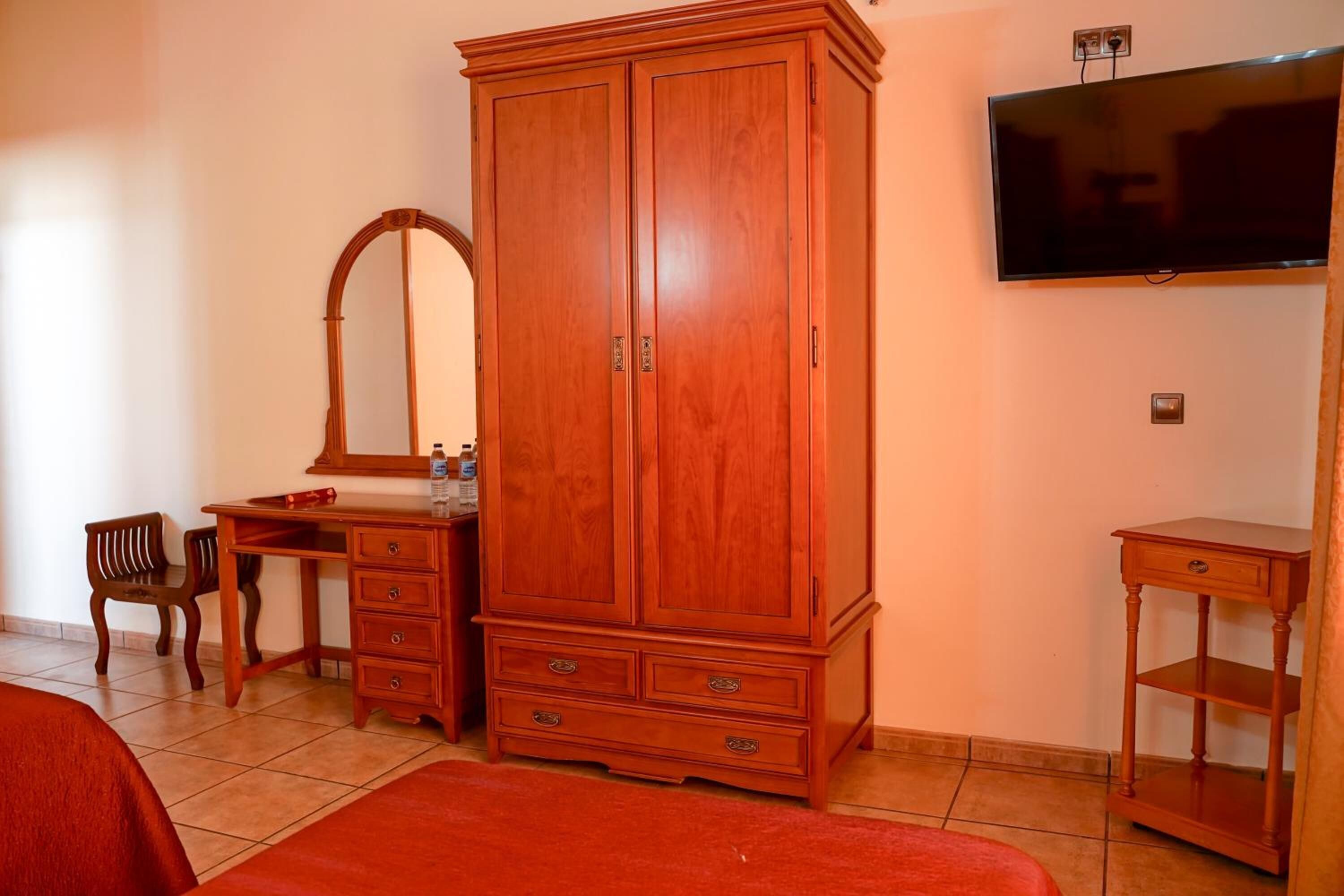 Une chambre avec une armoire en bois, un bureau, une chaise, une télévision et une petite table de chevet.