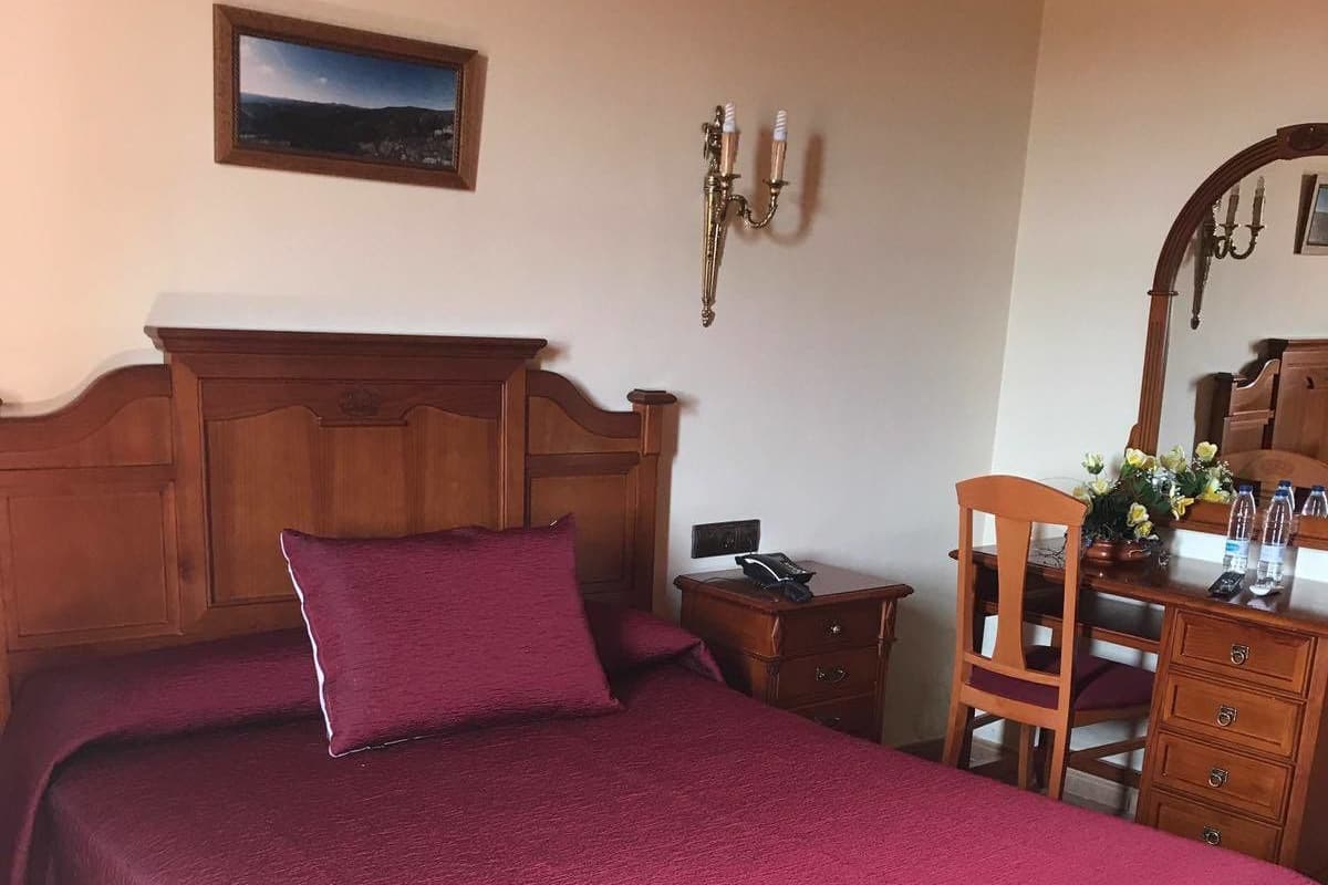 A bedroom with a wooden headboard, a large bed with a red bedspread, a nightstand, a wooden dresser, and a mirror.