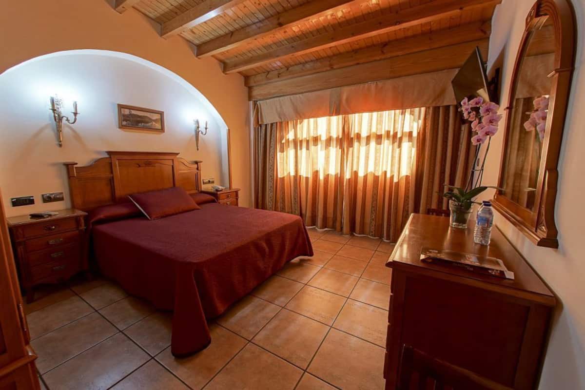 A bedroom with a large bed, wooden headboard, a nightstand, a dresser, a mirror, and a window with curtains.