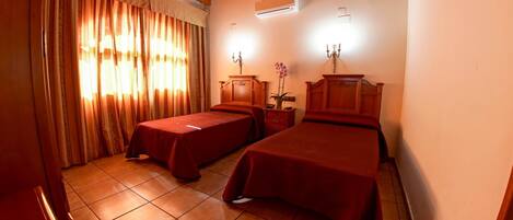 A hotel room with two beds, a wooden ceiling, a window with curtains, a nightstand with a lamp, and an air conditioning unit.