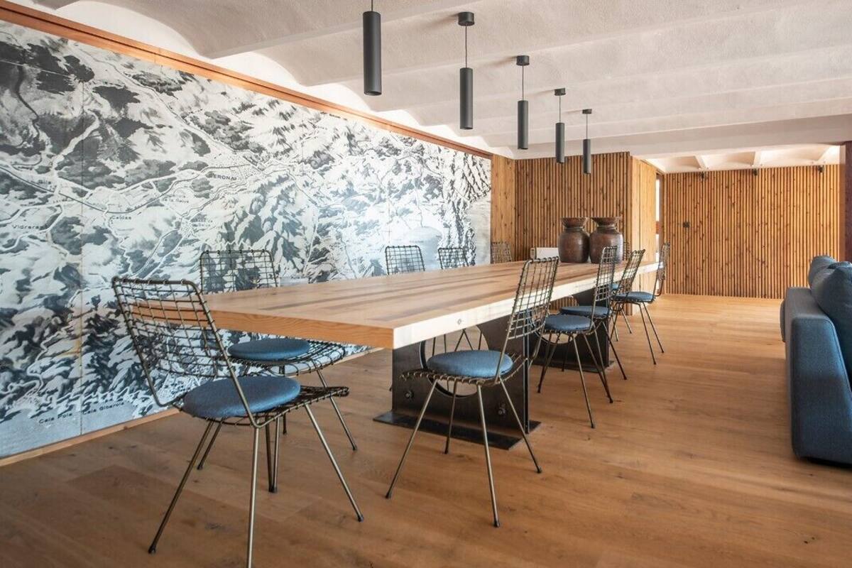 Uma área de refeições moderna com uma mesa de madeira comprida e cadeiras de estrutura de arame, um sofá azul e uma parede de madeira com um mural de mapa.