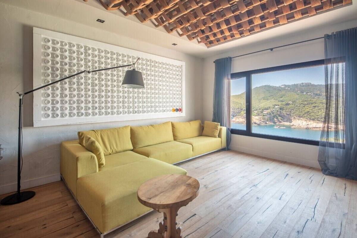 Quarto espaçoso com sofá amarelo, mesa de madeira e uma grande janela com vista para a paisagem costeira.