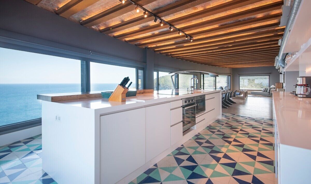 A modern kitchen with a large island, a view of the sea, and a geometric patterned floor.