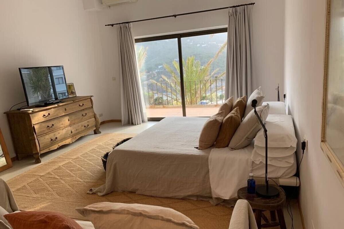 Quarto com cama, cômoda, luminária e vista para o exterior.