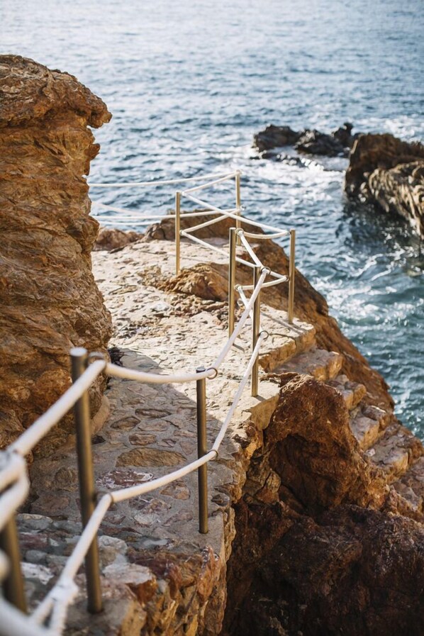 Escadaria de pedra descendo até uma orla rochosa, com guarda-corpo de metal.