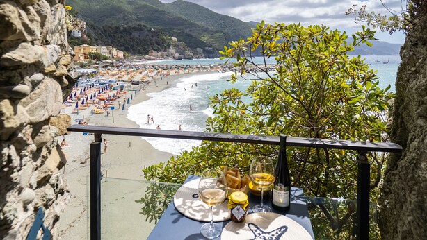 A table with a starfish mat, wine glasses, and a bottle, overlooking a beach with umbrellas and people. - La Casa del Gigante Luxury Collection (Monterosso al Mare)