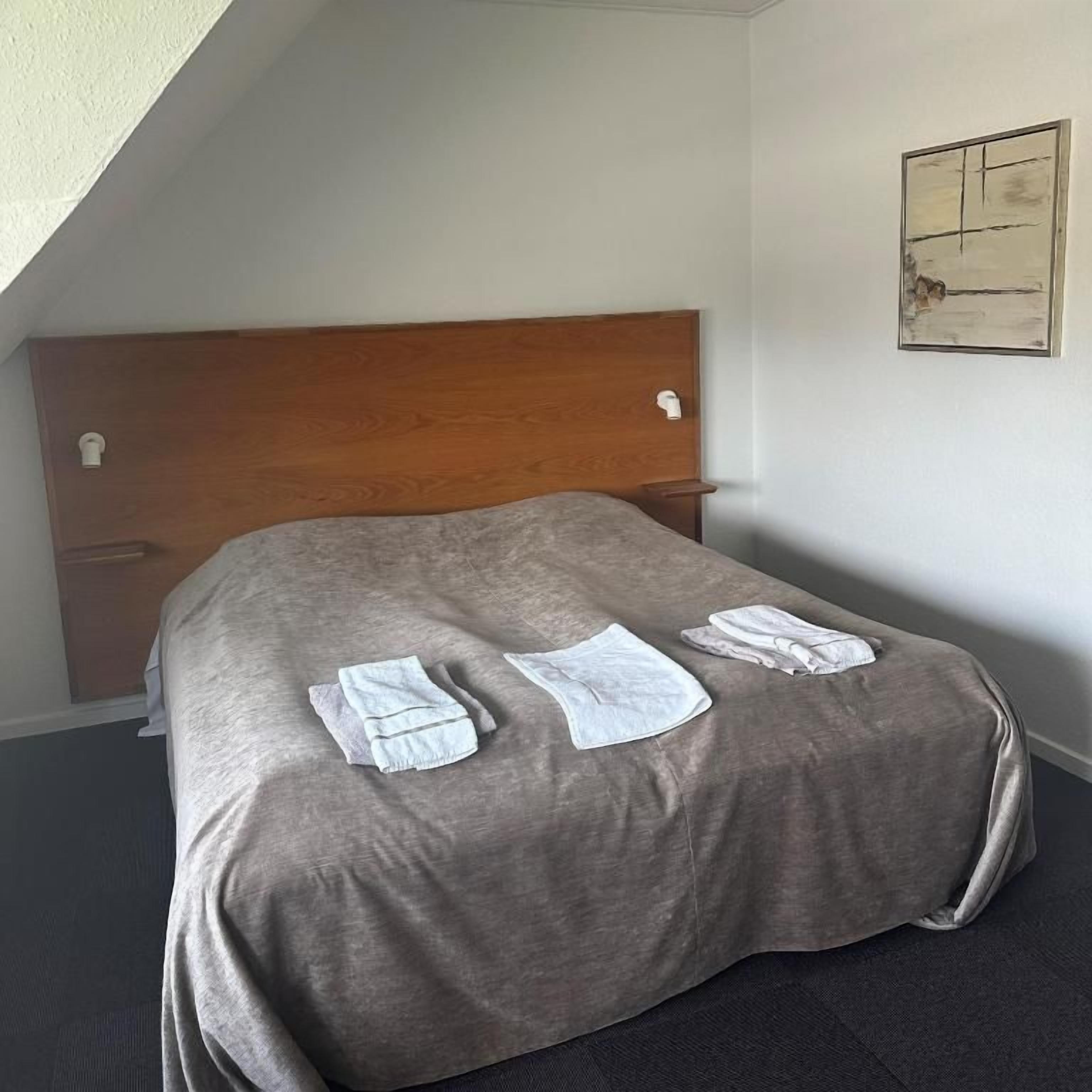 A single bed with a wooden headboard, a gray bedspread, and neatly folded white towels on the bed.