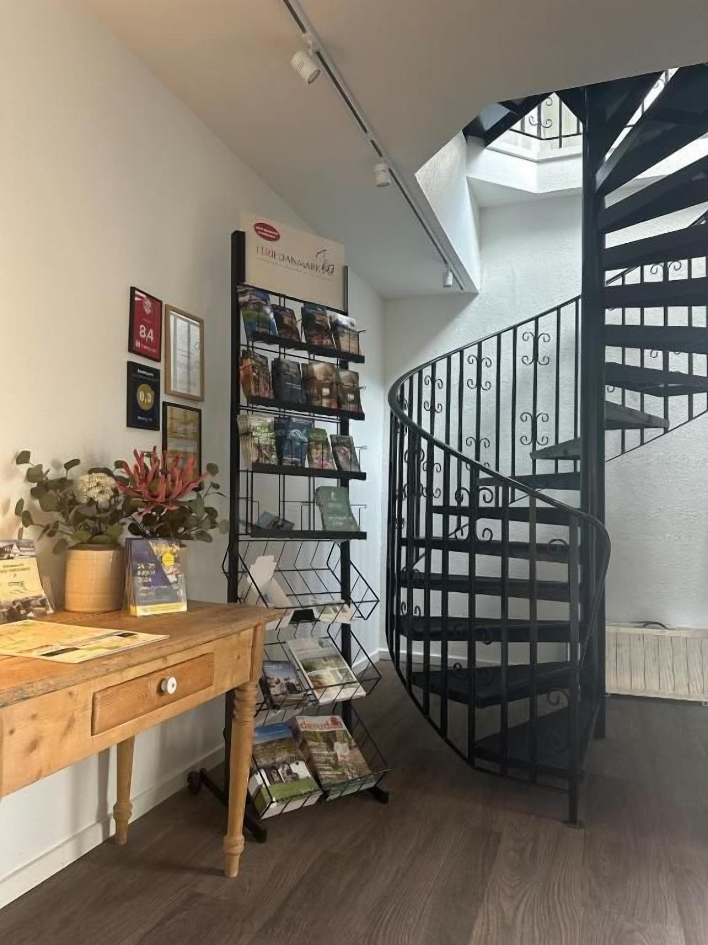 Un escalier en colimaçon avec une rampe en métal, un bureau en bois orné d’une plante, et une étagère où sont exposés des livres et des magazines.