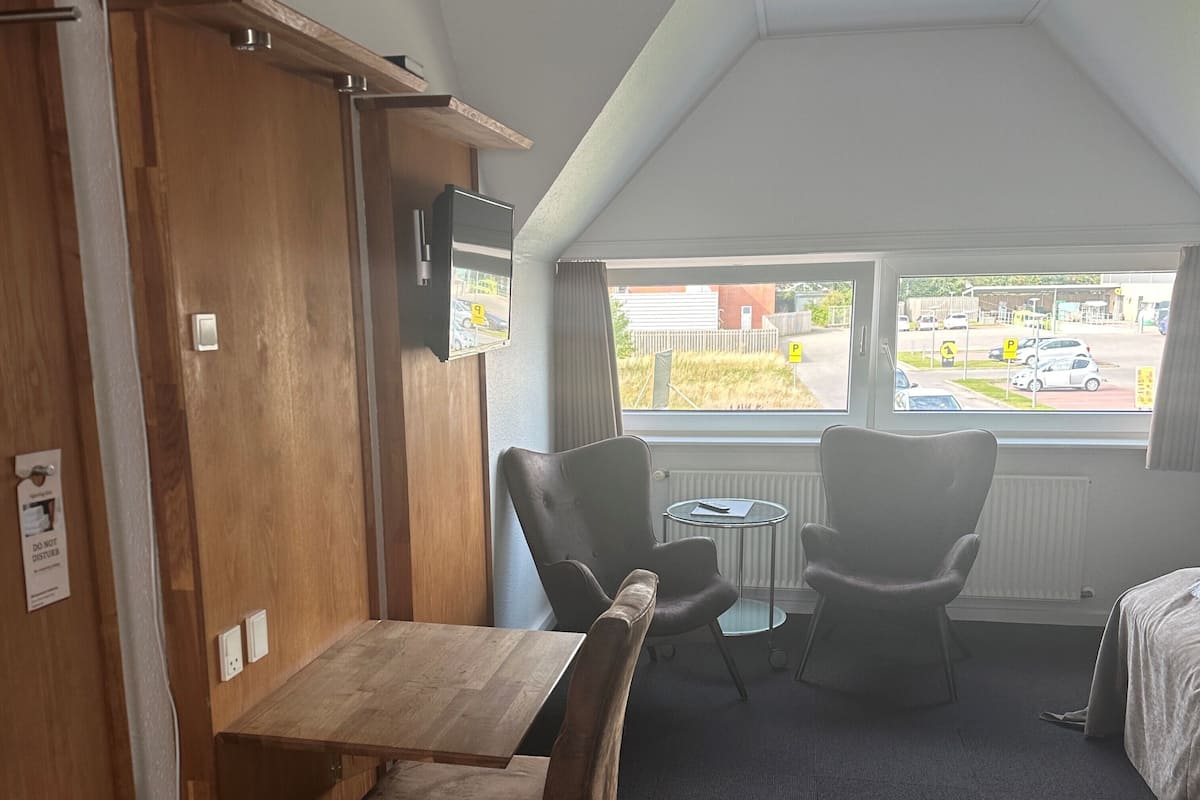 Hotellihuone, jossa on puinen vaatekaappi, pieni pöytä ja kaksi tuolia, ikkuna, josta avautuu näkymä parkkeerattuihin autoihin ja rakennuksiin, sekä patteri ikkunan alla.