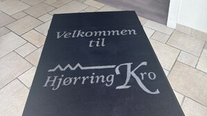 A welcome mat with "Velkommen til Hjørring Kro" text, leading to a hallway with a dining area and a lamp. - Hjørring Kro (Hjørring)