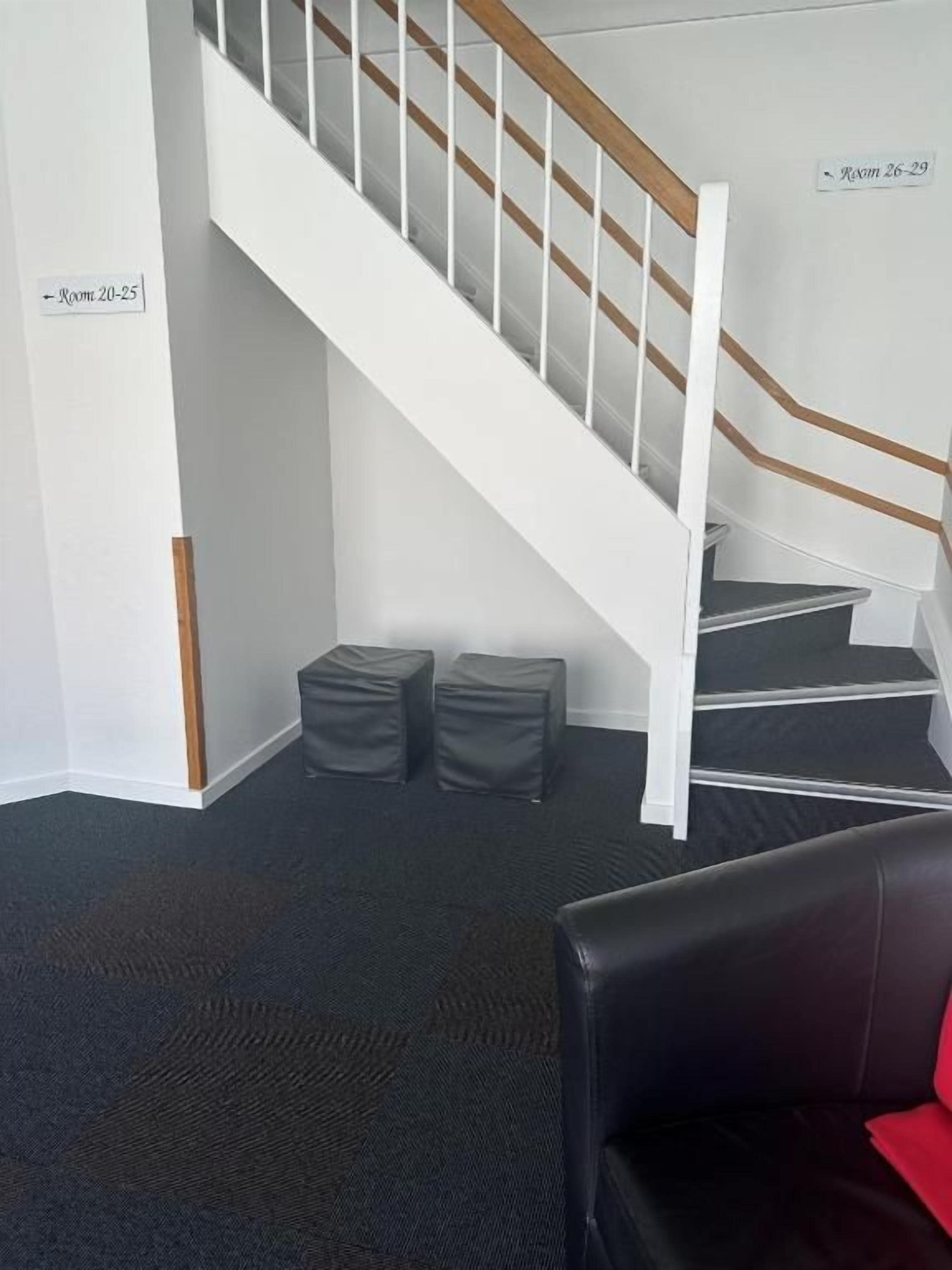 A staircase with white railings and a black carpeted floor. There are two black cushioned stools and a black leather sofa with a red cushion.