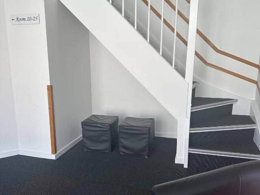 A staircase with white railings and a black carpeted floor. There are two black cushioned stools and a black leather sofa with a red cushion.