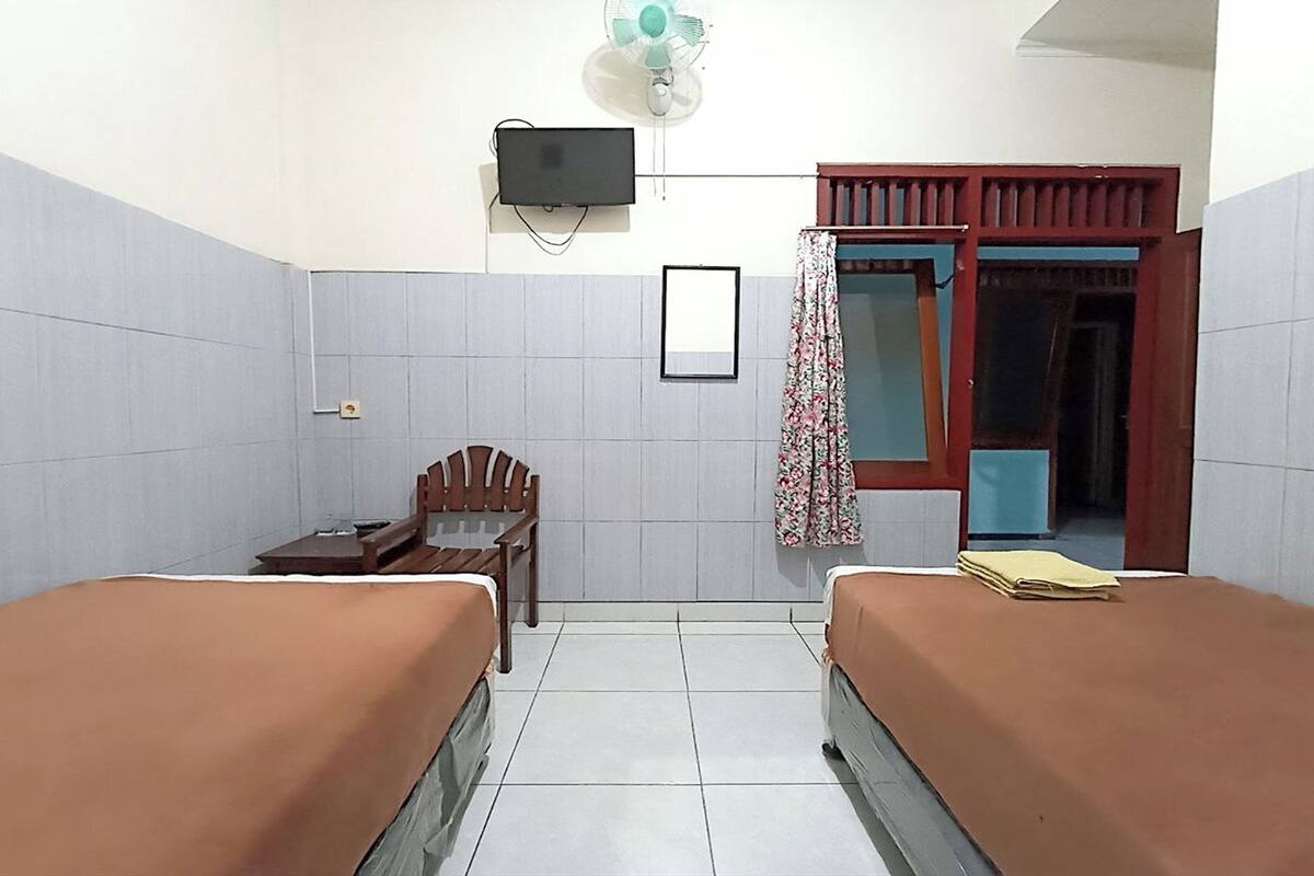 A small, clean room with two beds, a desk, a chair, a TV, and a window with a curtain.