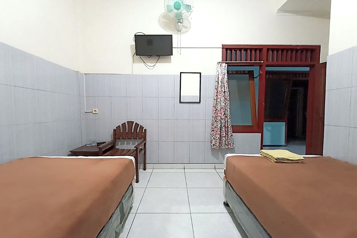 A small, clean room with two beds, a desk, a chair, a TV, and a window with a curtain.
