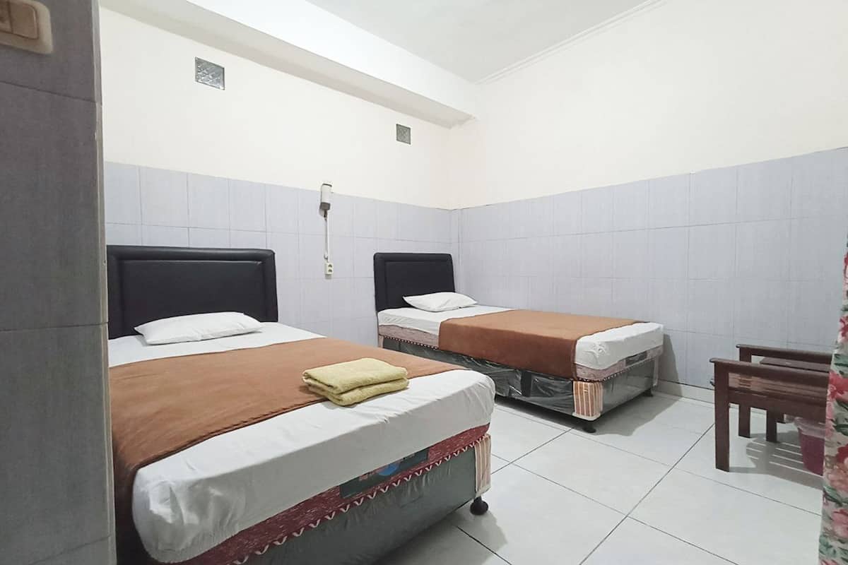 A room with two beds, each with a pillow and a blanket. There is a chair and a small table in the corner.