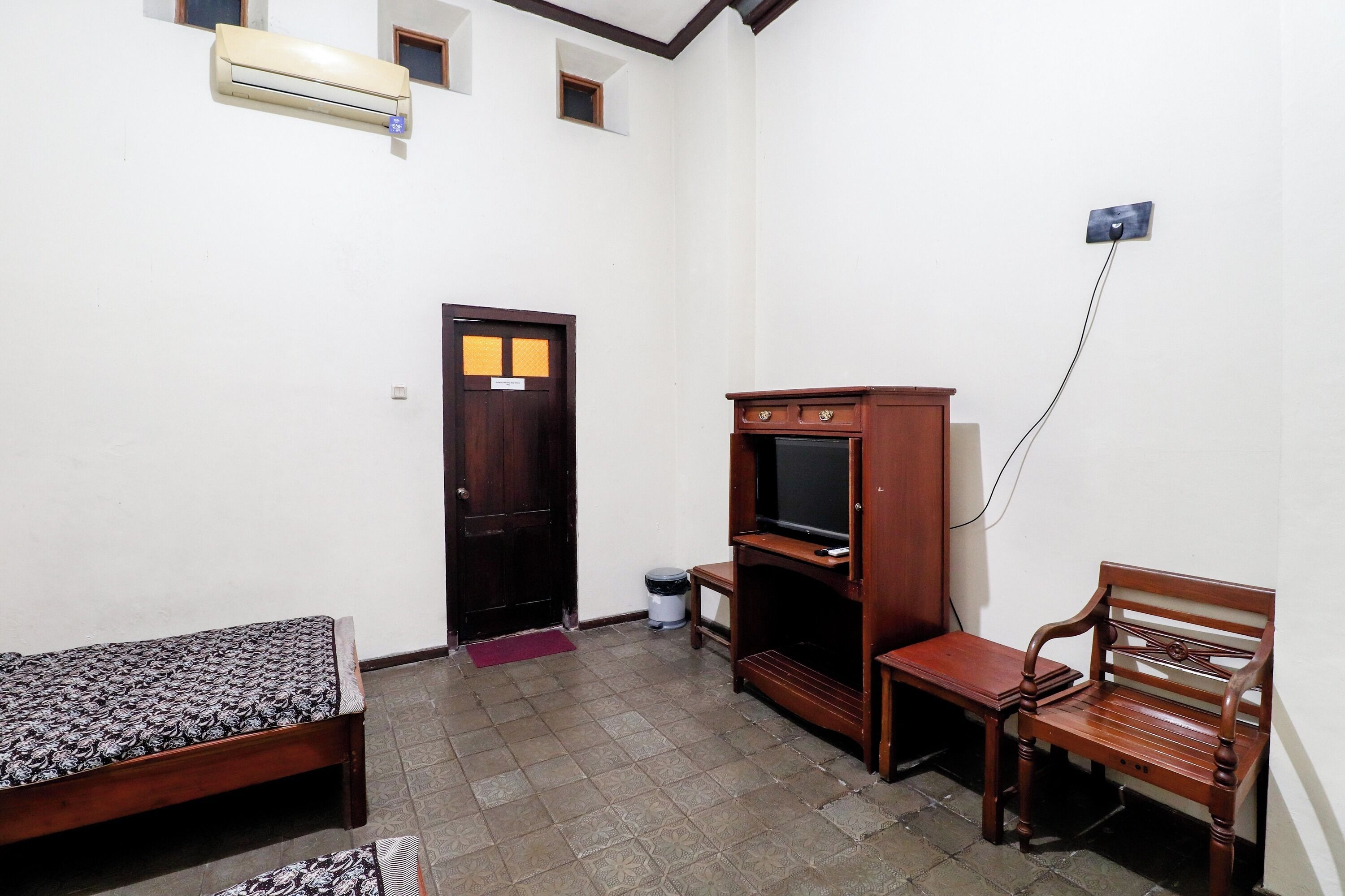 A small, dimly lit room with a single bed, a wooden bench, a television, and a small trash bin.