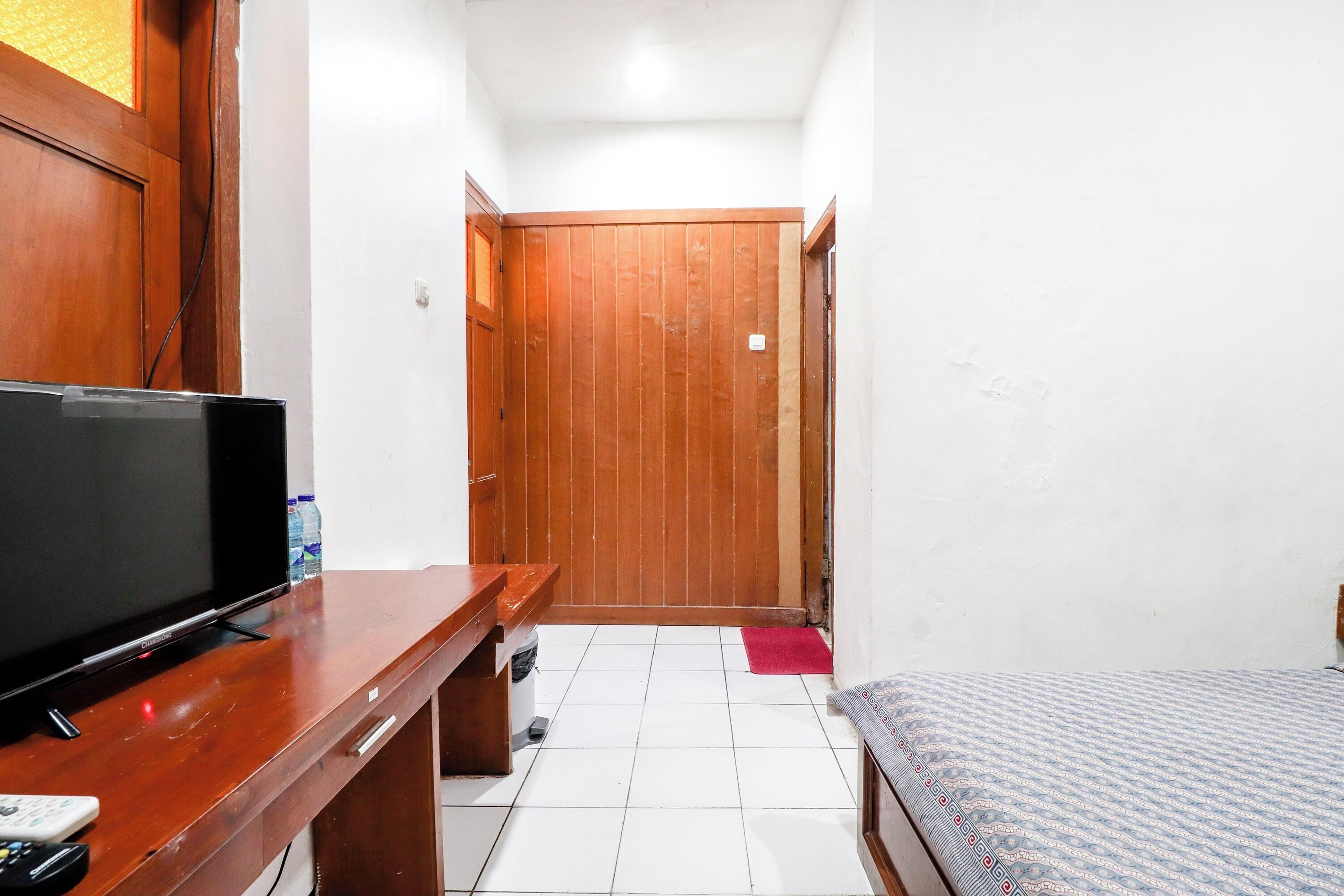 A small, compact room with a wooden desk, a television, a bed, and a door leading to another room.