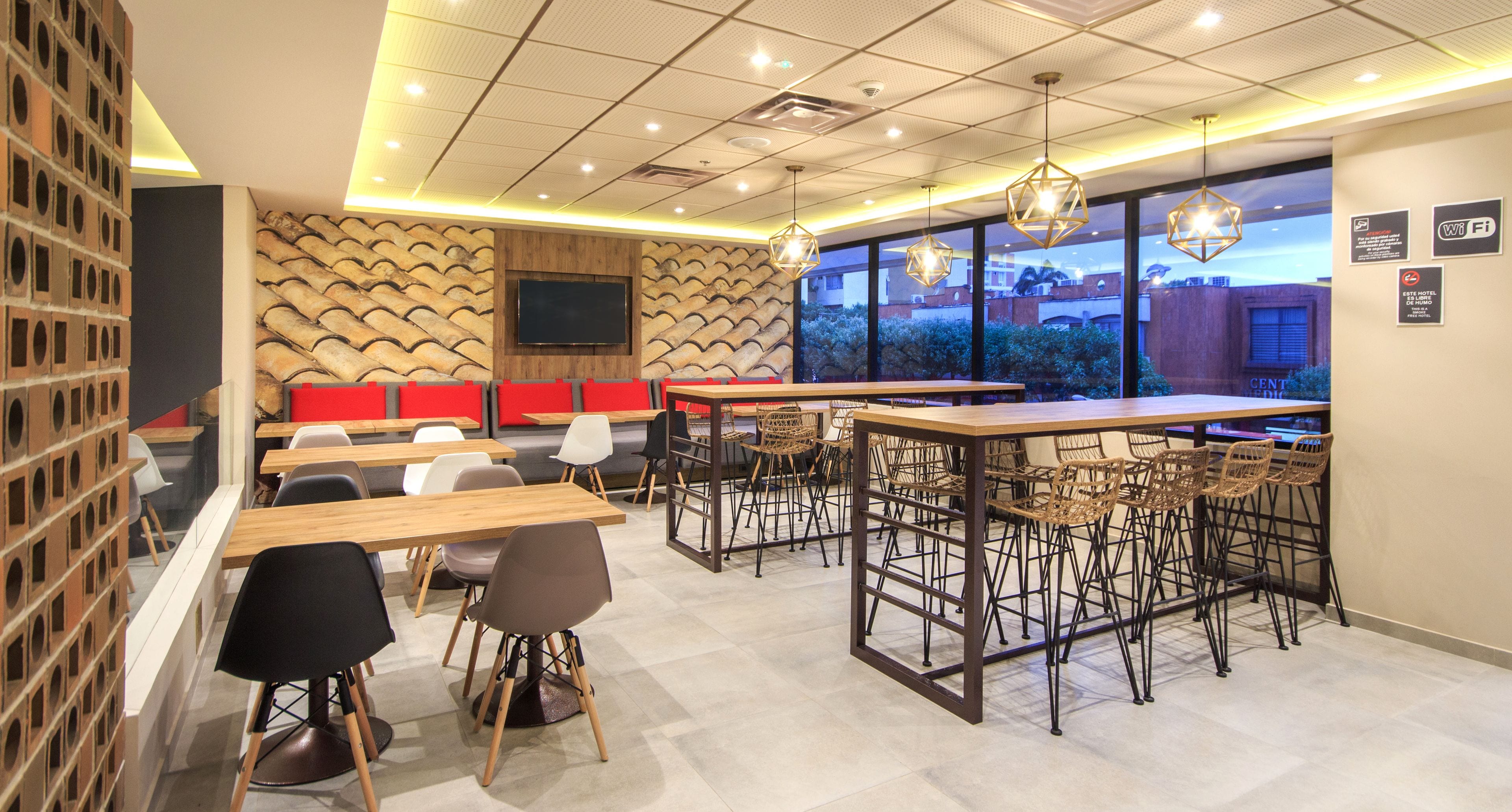 Un café moderno con mesas y sillas de madera, un televisor montado en la pared y un gran ventanal con vistas a la ciudad.