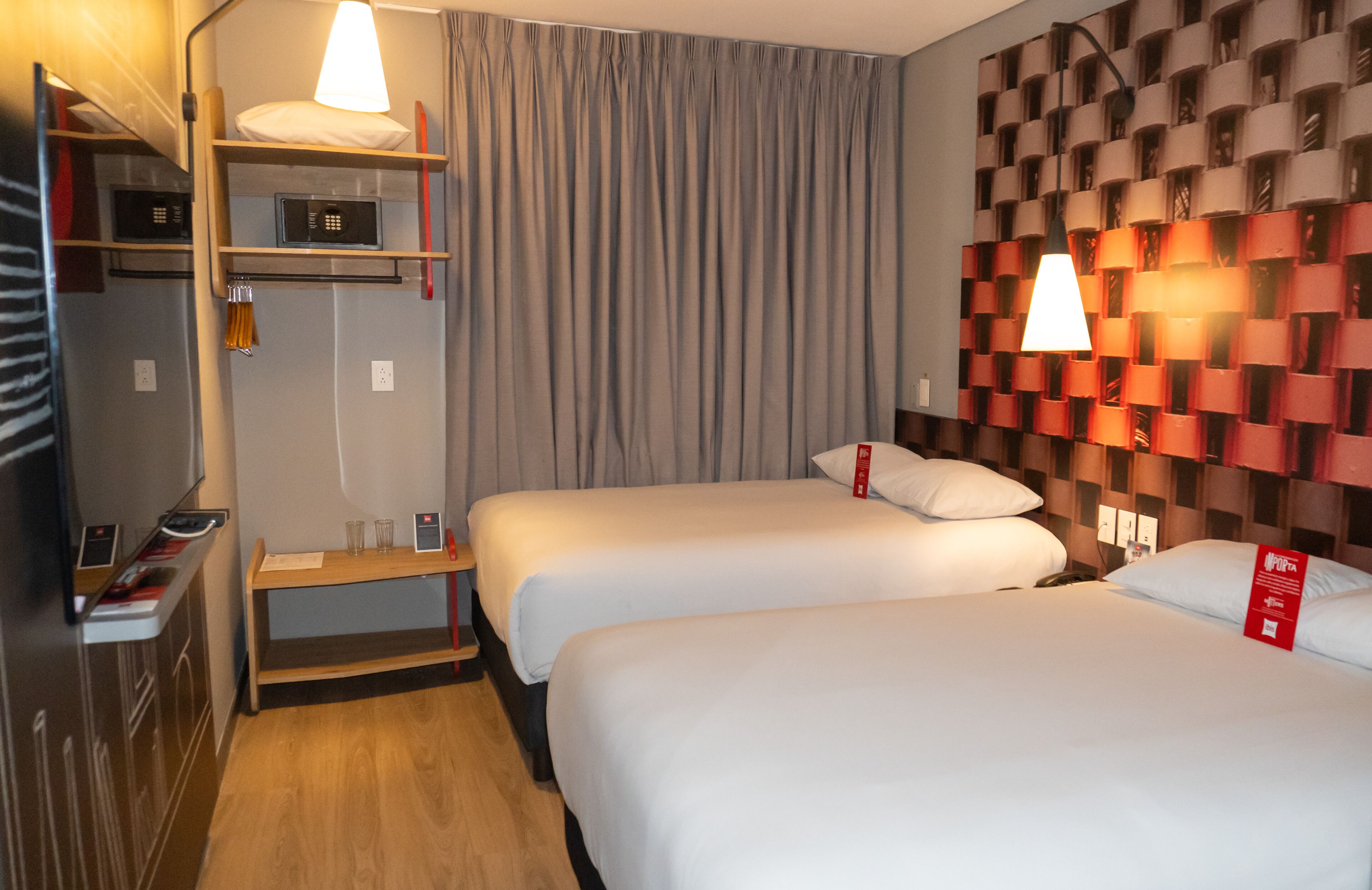 A hotel room with two beds, a desk with a lamp, a wardrobe, and a wall with a unique pattern.