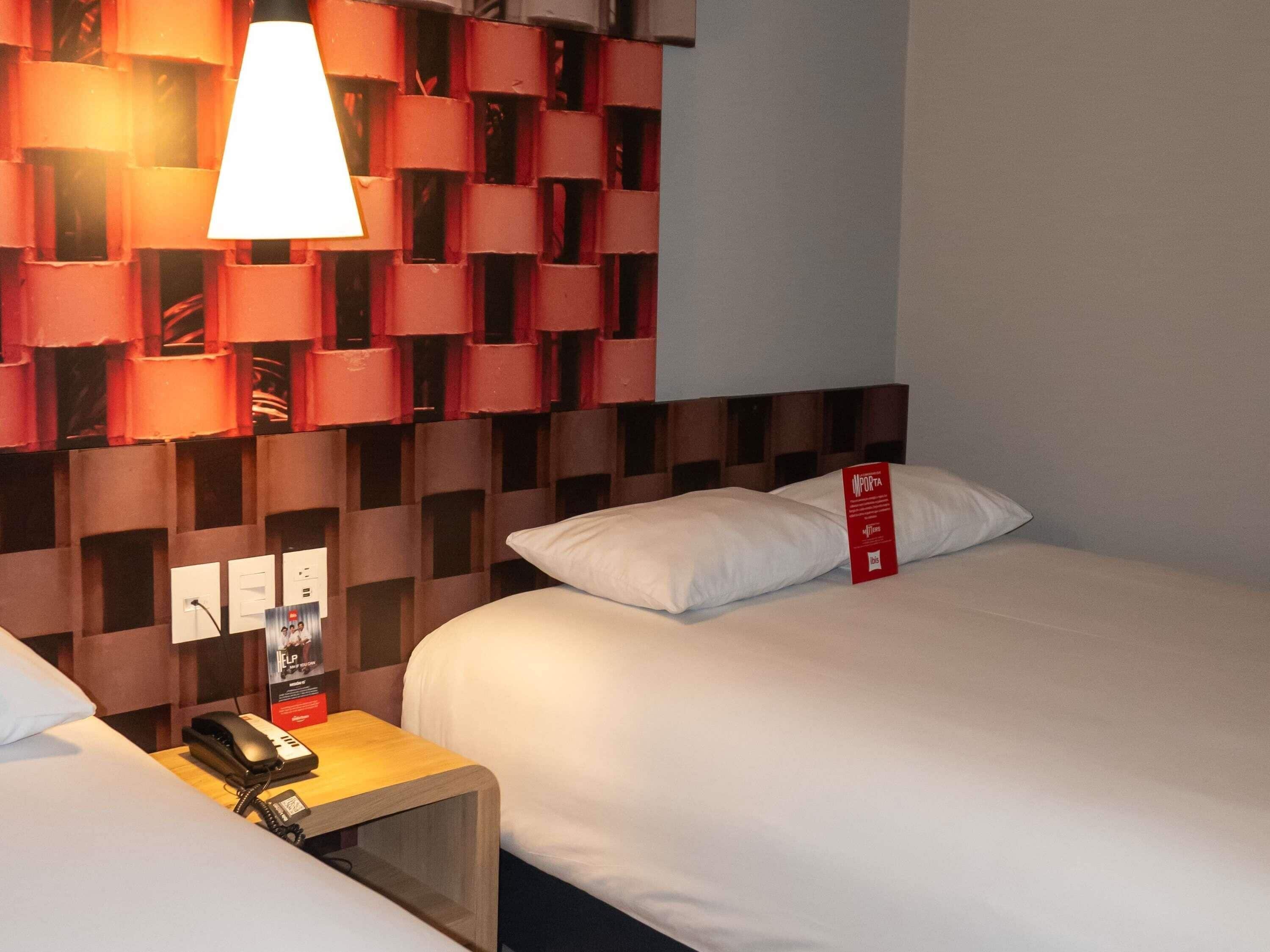 Een hotelkamer met twee bedden, een nachtkastje met een telefoon, een rood bord en een muur met een geometrisch patroon.