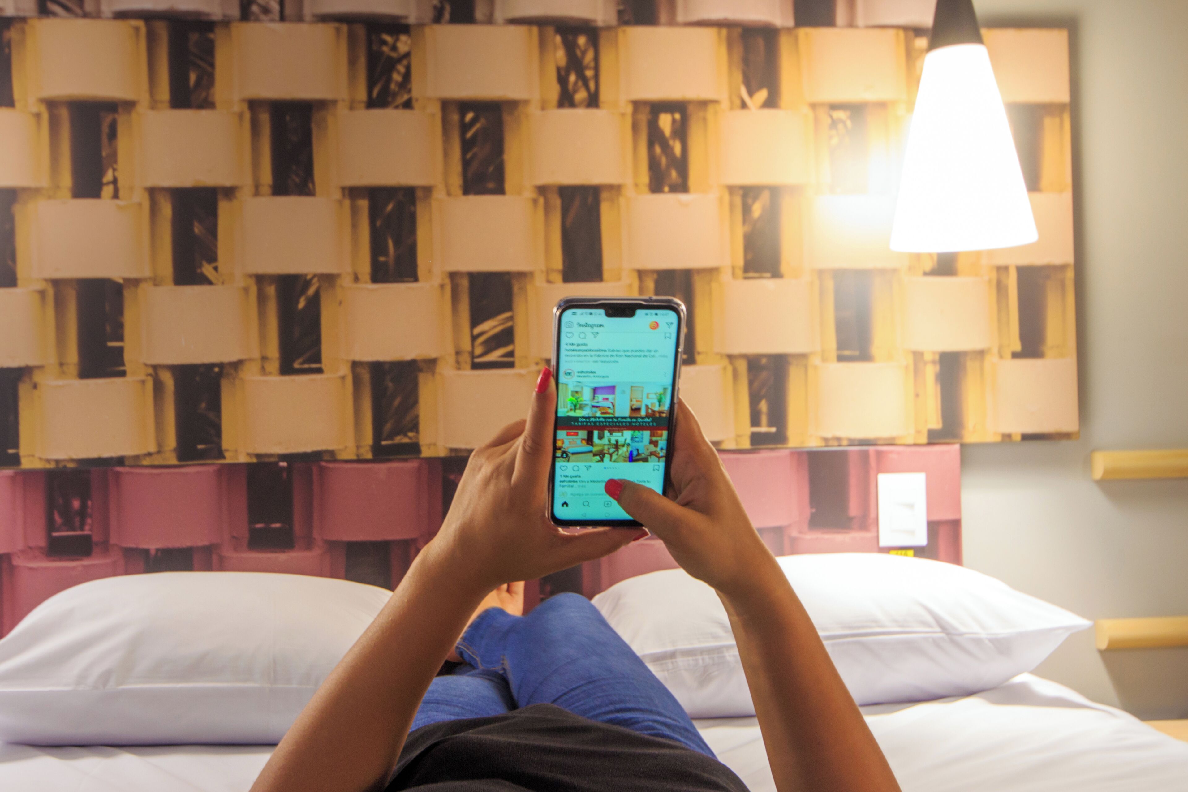 Una persona acostada en una cama con un teléfono inteligente, un papel tapiz estampado y una lámpara colgante.