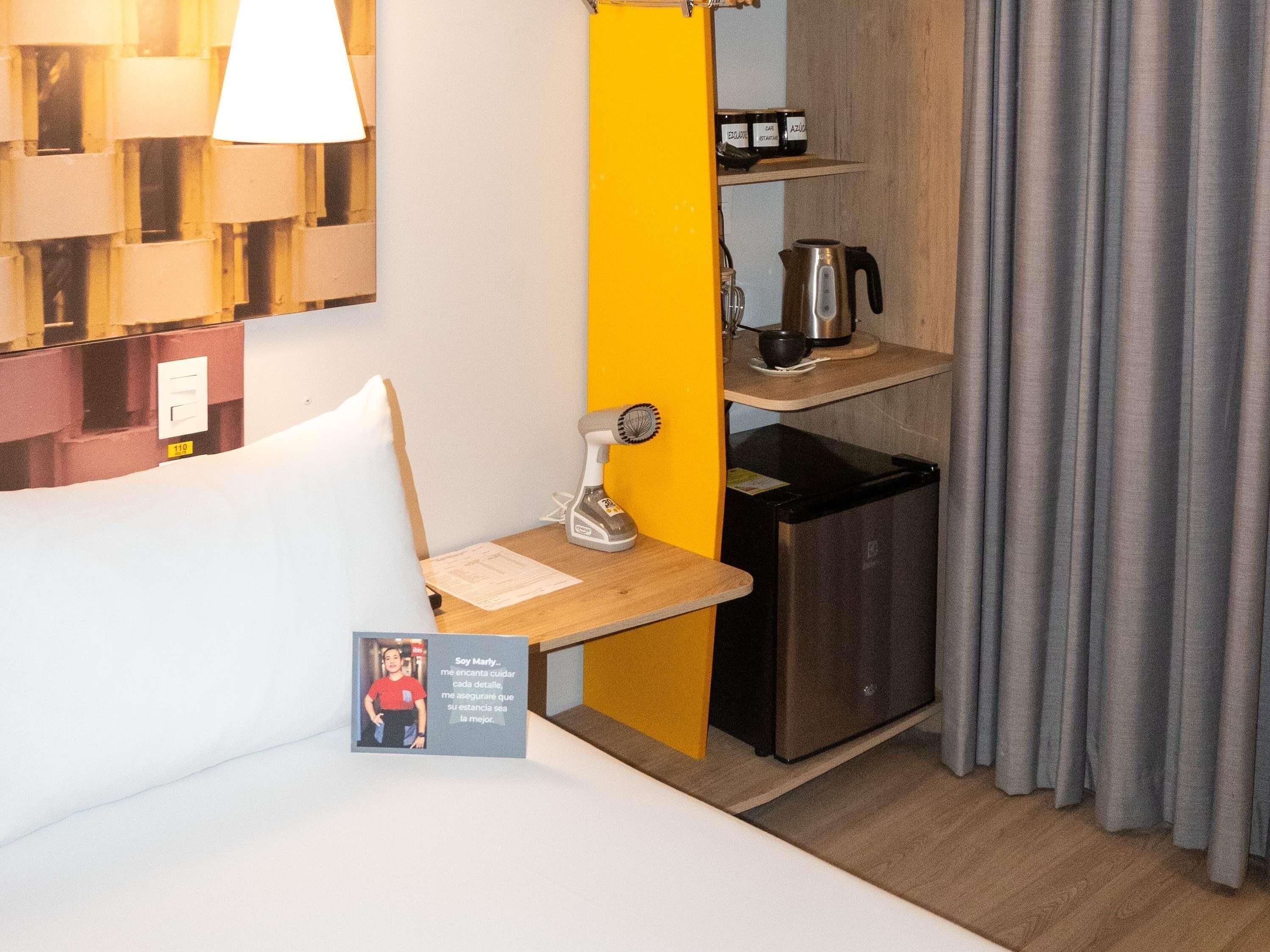 A compact hotel room with a bed, a small desk with a lamp, a coffee maker, and a mini-fridge.