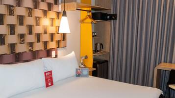 Una habitación de hotel con una cama, una lámpara colgante, una mesita, una silla, un perchero y un estante en la pared con una tetera.