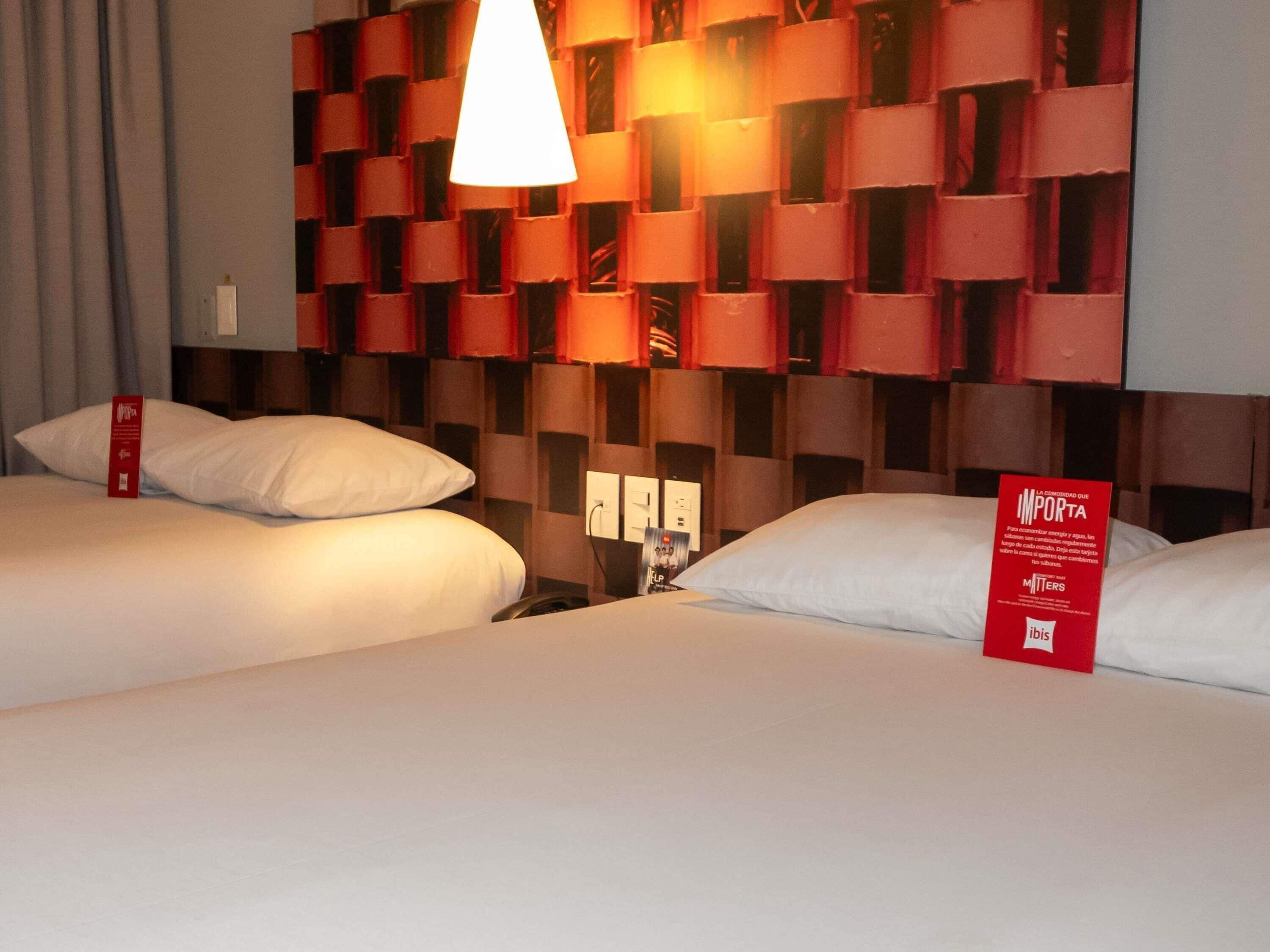A hotel room with two beds, a red wall with a geometric pattern, a white hanging light fixture, and a red sign on the wall.