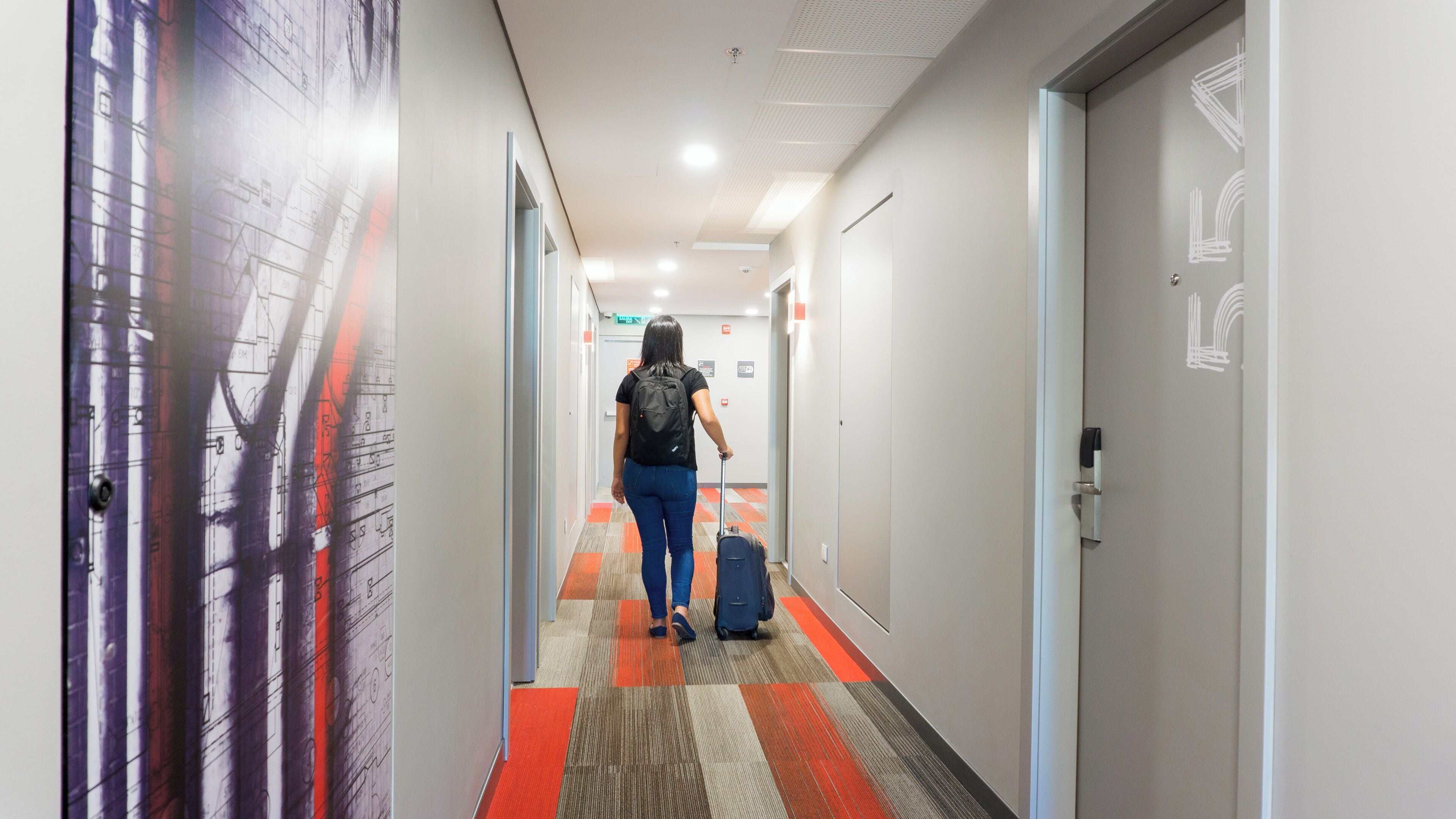A hallway with a patterned carpet, a person with a suitcase, and doors with a simple design.