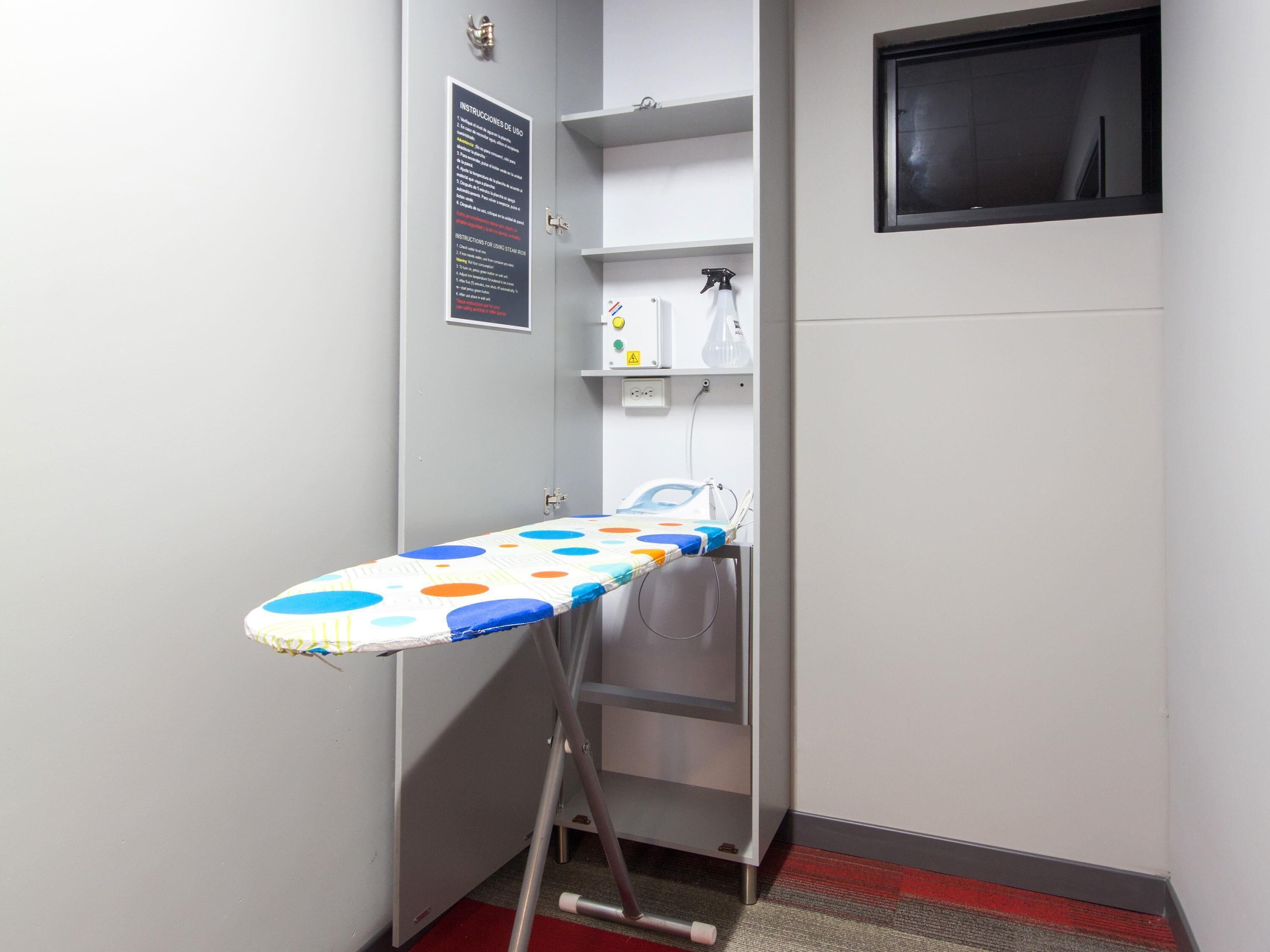 A corner room with a wall-mounted ironing board, a small shelf with cleaning supplies, and a door with a sign.