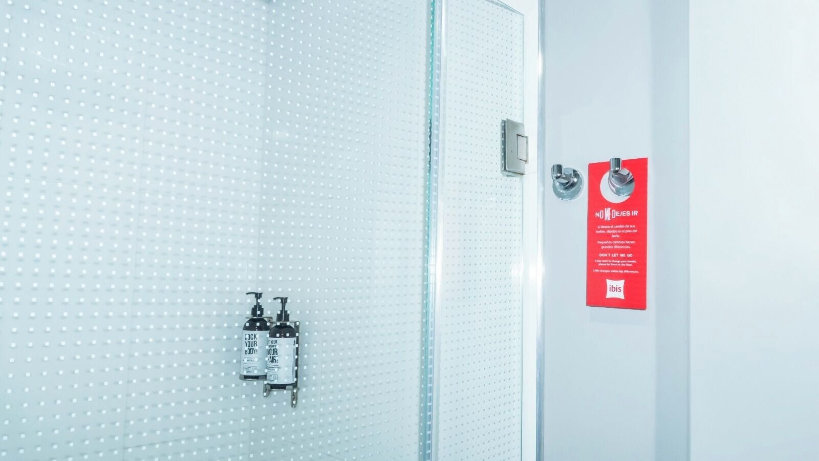 Un baño con pared de azulejos blancos, zona de ducha y un cartel informativo rojo.