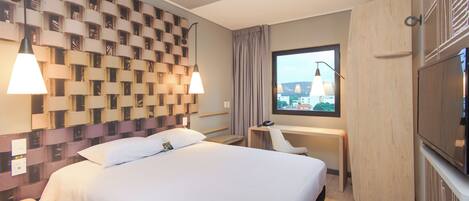 Ett hotellrum med en stor säng, ett skrivbord, en stol, en TV och ett fönster med utsikt över staden.