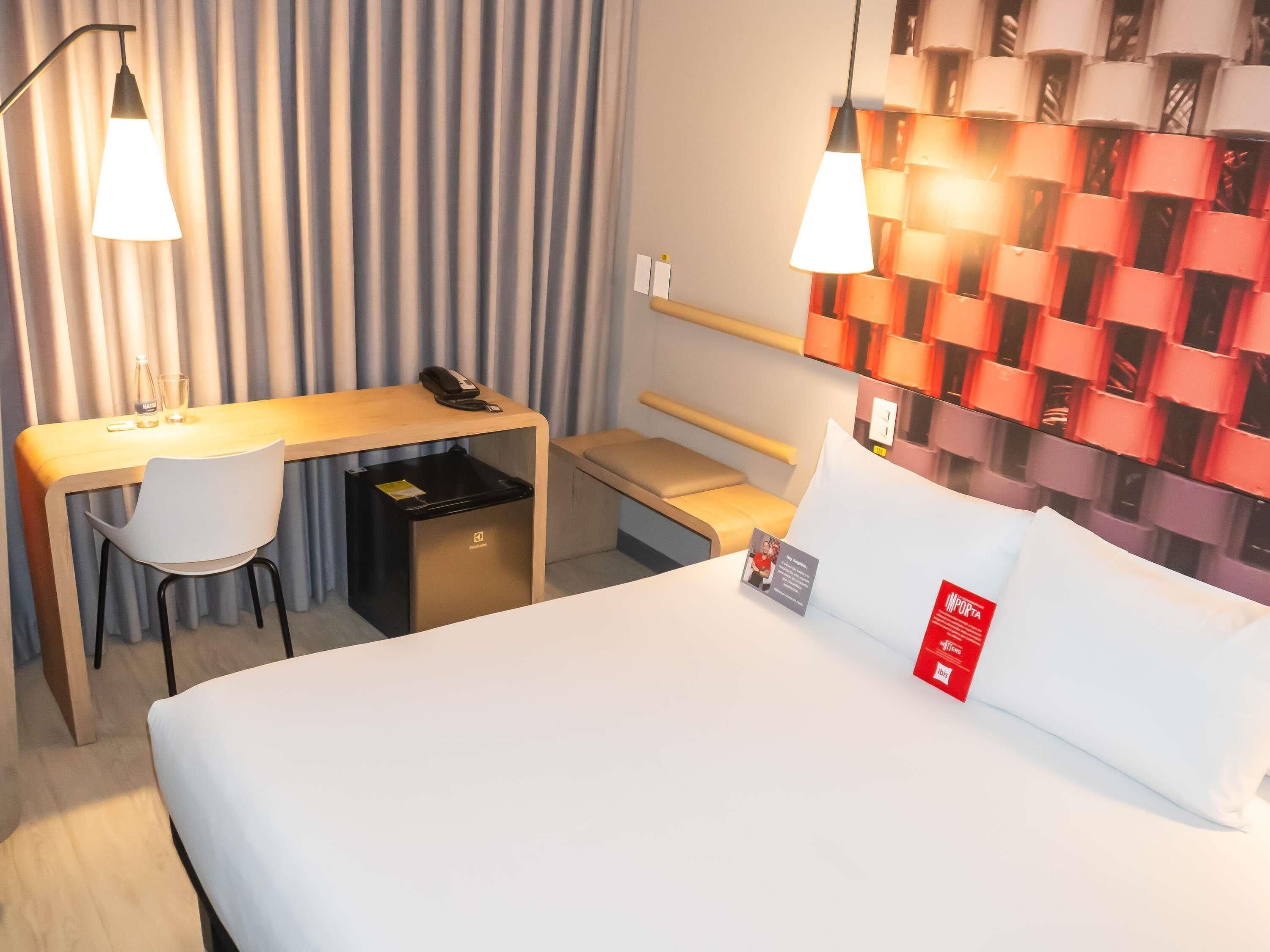 Et hotelværelse med seng, skrivebord, stol, skraldespand og en lampe ophængt på væggen.
