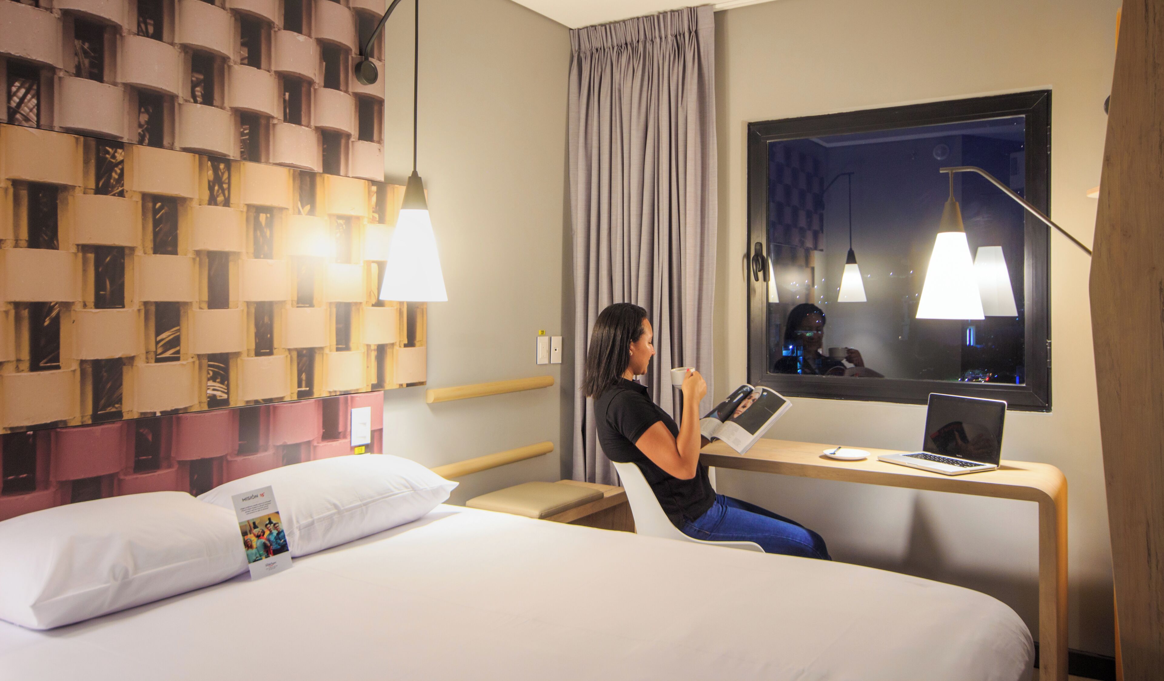 A hotel room with a bed, a desk with a laptop, a mirror, and a wall with a patterned design.