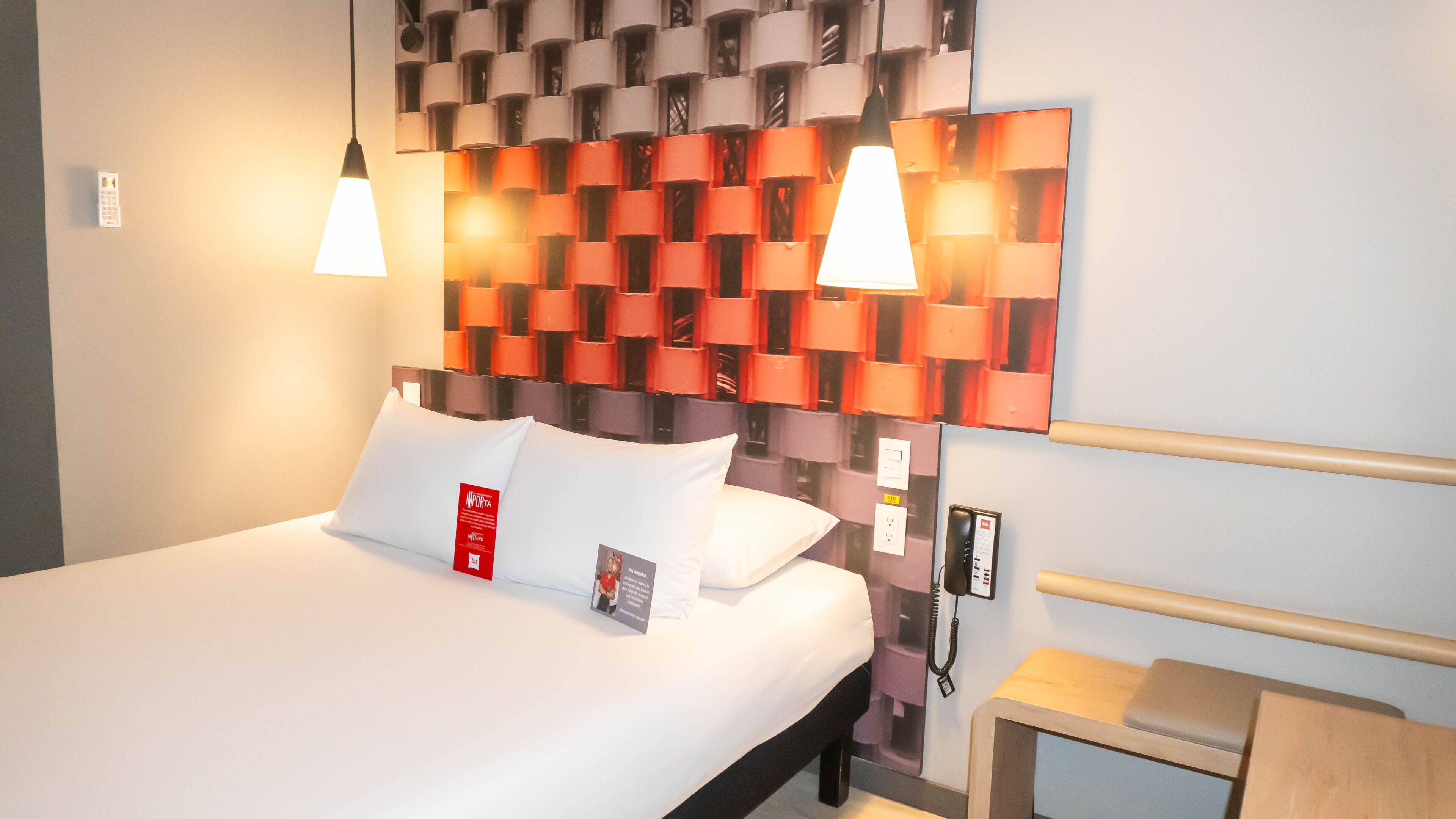 A hotel room with a neatly made bed, two pendant lights, a modern wall headboard, and a bedside table with a lamp.