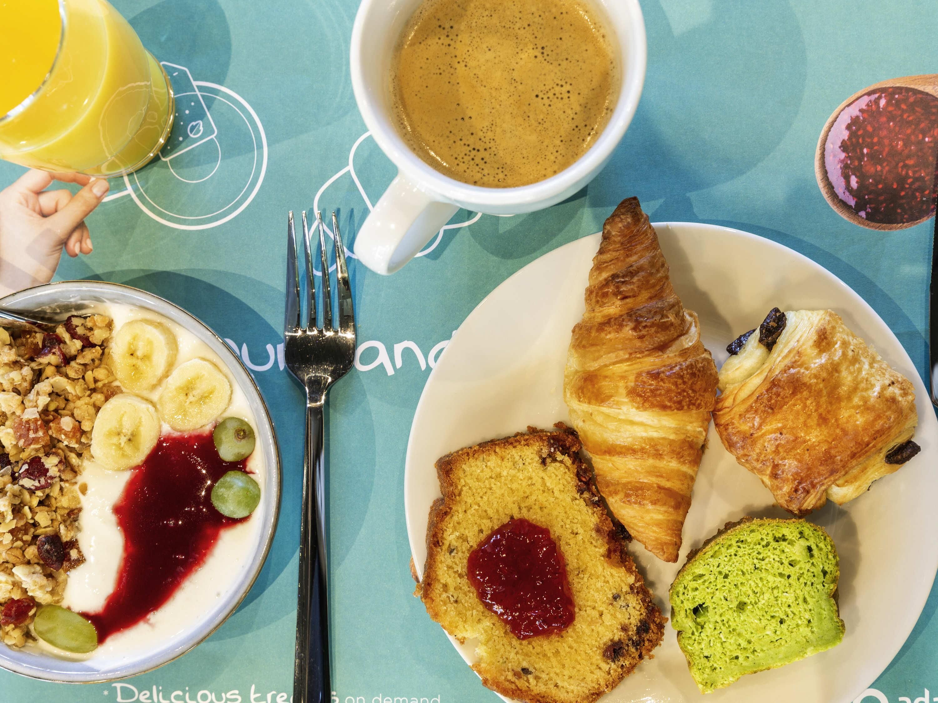 Un desayuno con una fuente de cruasanes, una porción de pastel con mermelada, un tazón de yogur con fruta y una taza de café.