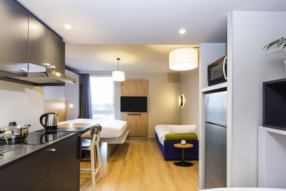 Une cuisine moderne équipée d’une cuisinière, d’une bouilloire et de placards, un coin repas avec une table et des chaises, un petit coin salon avec un canapé et une table d’appoint, et un téléviseur fixé au mur.