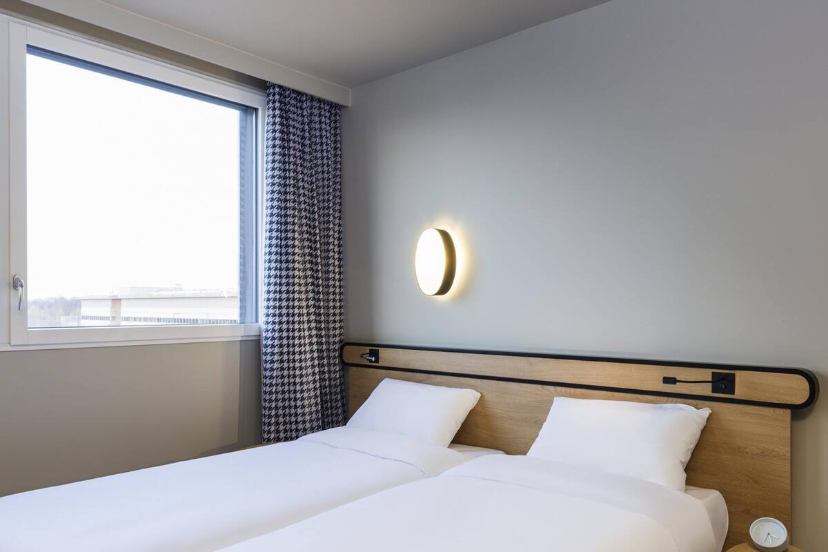 Une chambre d’hôtel avec un lit, une table de chevet avec une horloge, une lampe fixée au mur et une fenêtre avec des rideaux.